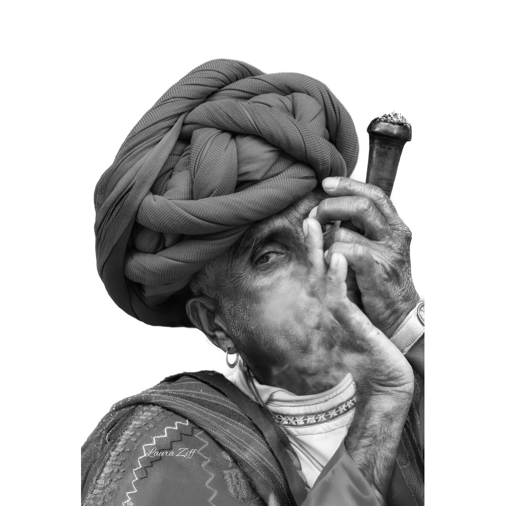 Black and white photo of a Rabari wearing a turban made of cloth, smoking a chillum, with smoke coming from their mouth and cigarette. The person has earrings, a watch, and a patterned collar on their shirt.