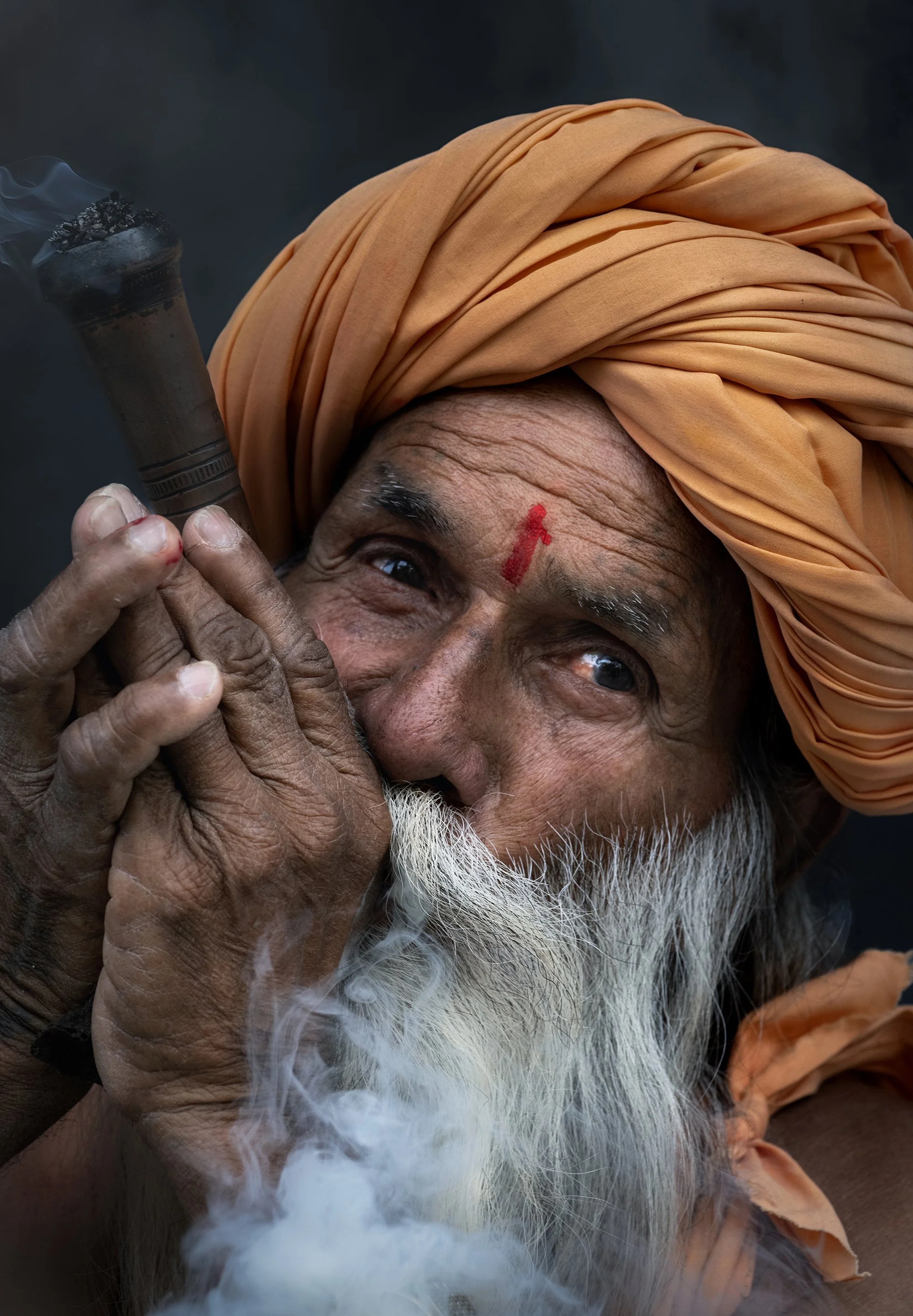 An elderly Indian man with a long white beard and orange turban, holding a smoking pipe close to his face, looking at the camera with a gentle smile.