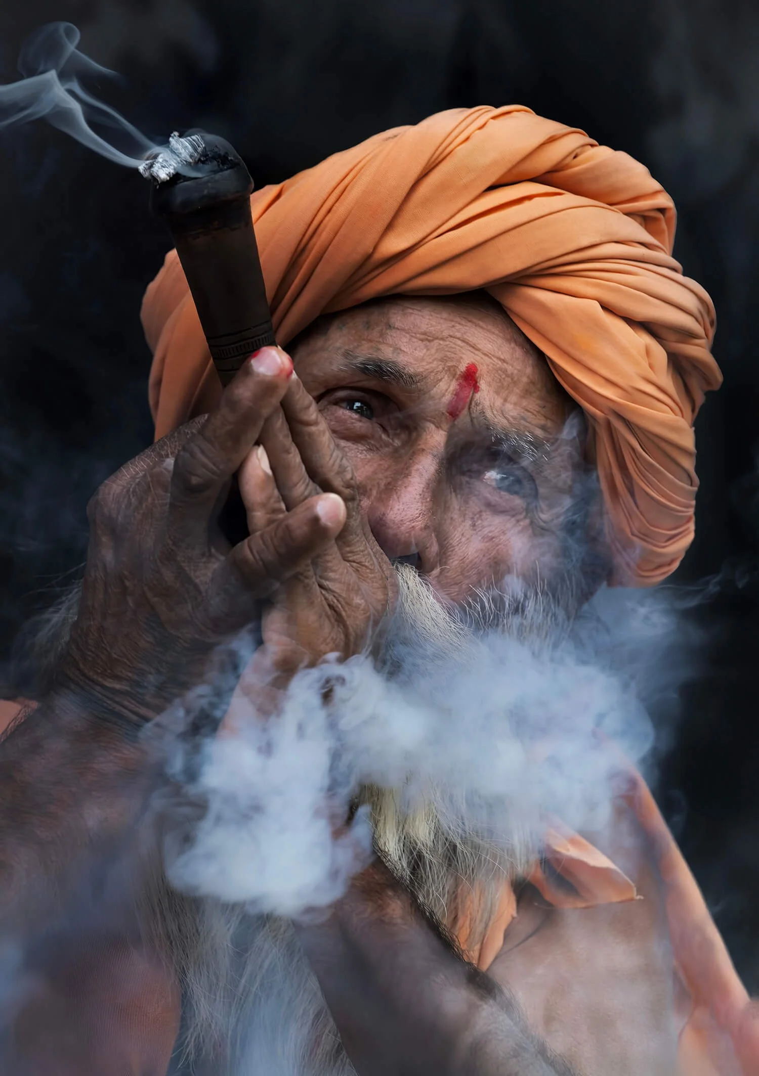 A Rabari herdsman wearing an orange turban smoking a chillum. It is their tradition at the end of the work day to get together and pass the pipe.  The tobacco is insanely strong.  Photographed in Jawai, india.