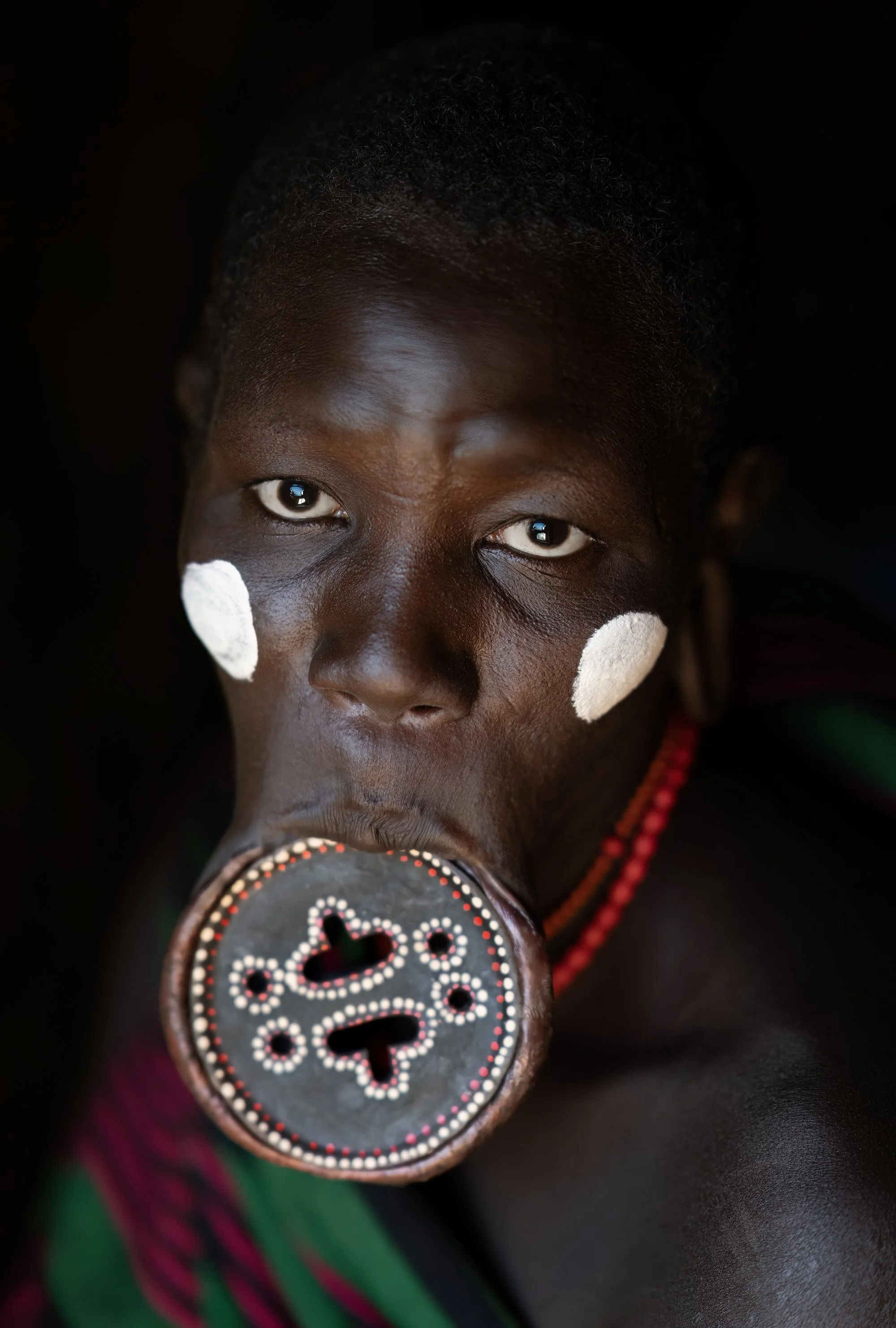A person with traditional face paint and jewelry, holding a decorated object in their mouth, looking directly at the camera.