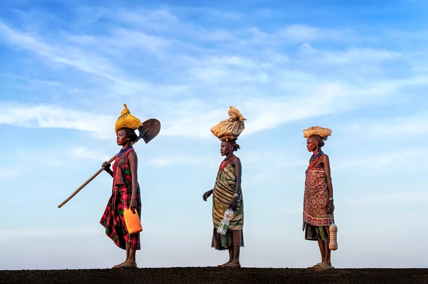 I could not have planned this photo if I tried.  Three women carrying items on their heads and holding bottles, standing in a line walking home from a long day of work.  Photograph taken in the Omo Valley, Ethiopia. 