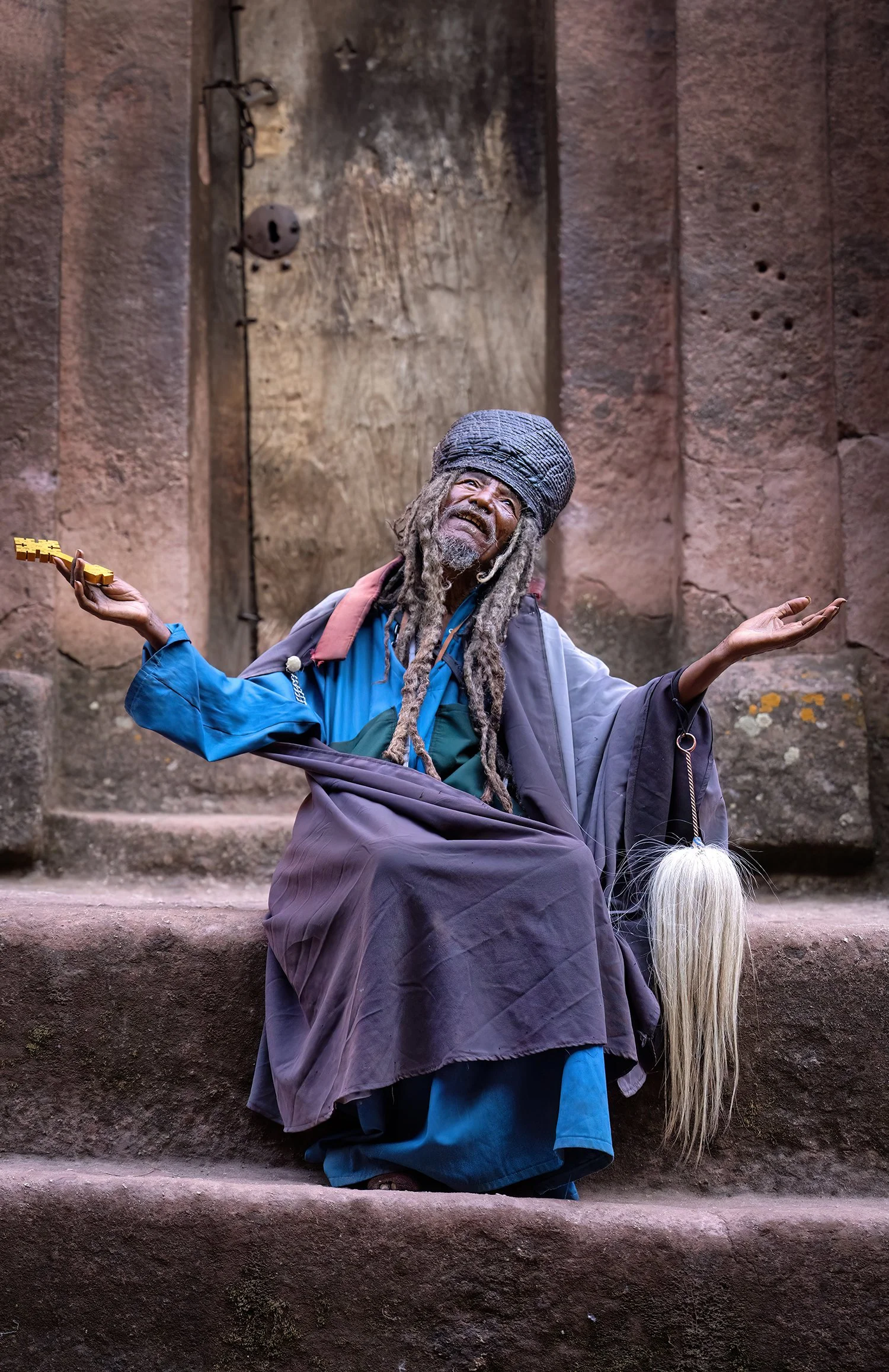 A person dressed as a traditional Rastafarian with dreadlocks, wearing colorful clothing and a headwrap, sitting on stone steps with a serious expression, with arms extended outwards and palms up.
