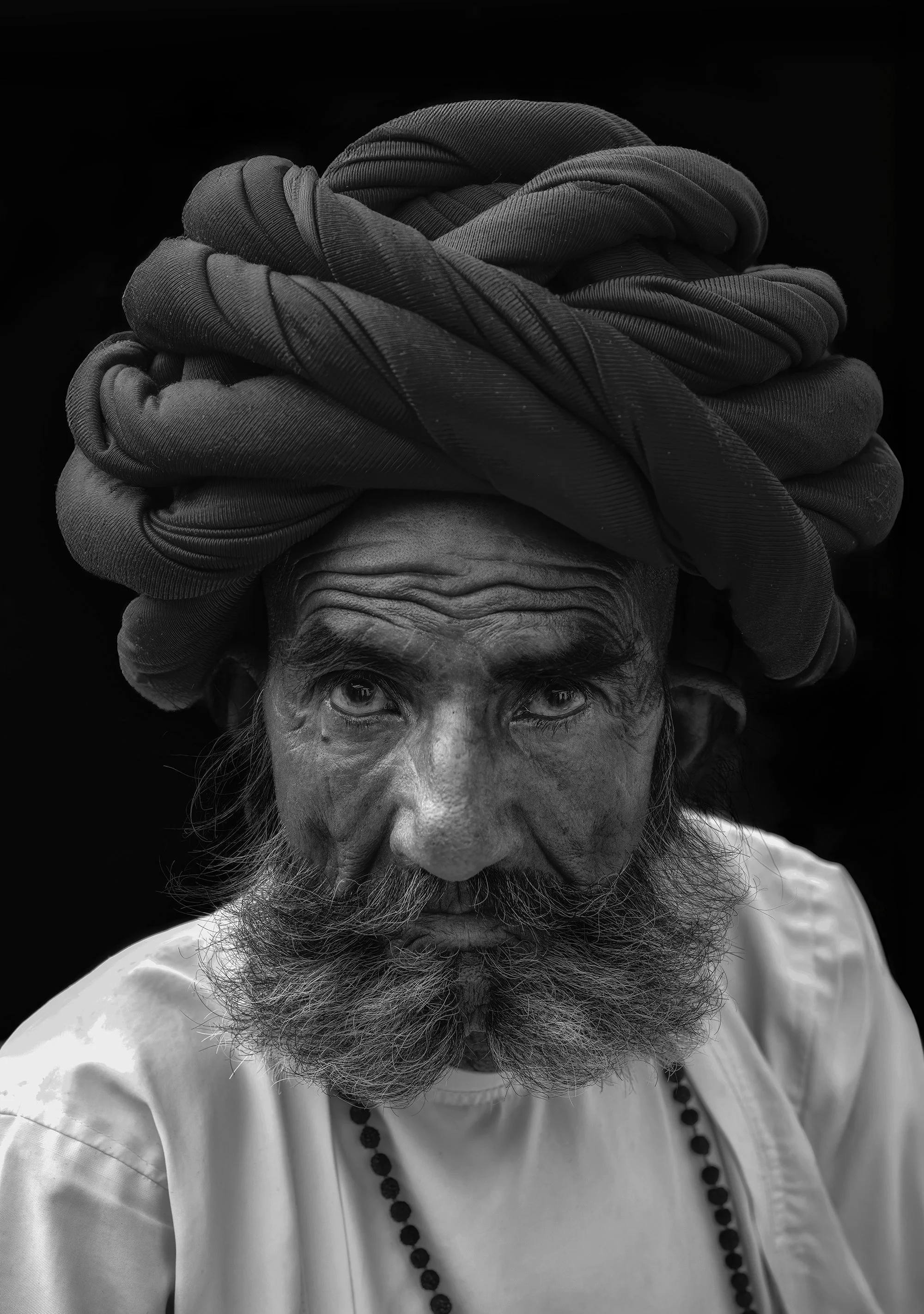 A black and white portrait of an elderly man with a deeply wrinkled face, a thick beard, wearing a traditional turban and a white shirt with a beaded necklace.