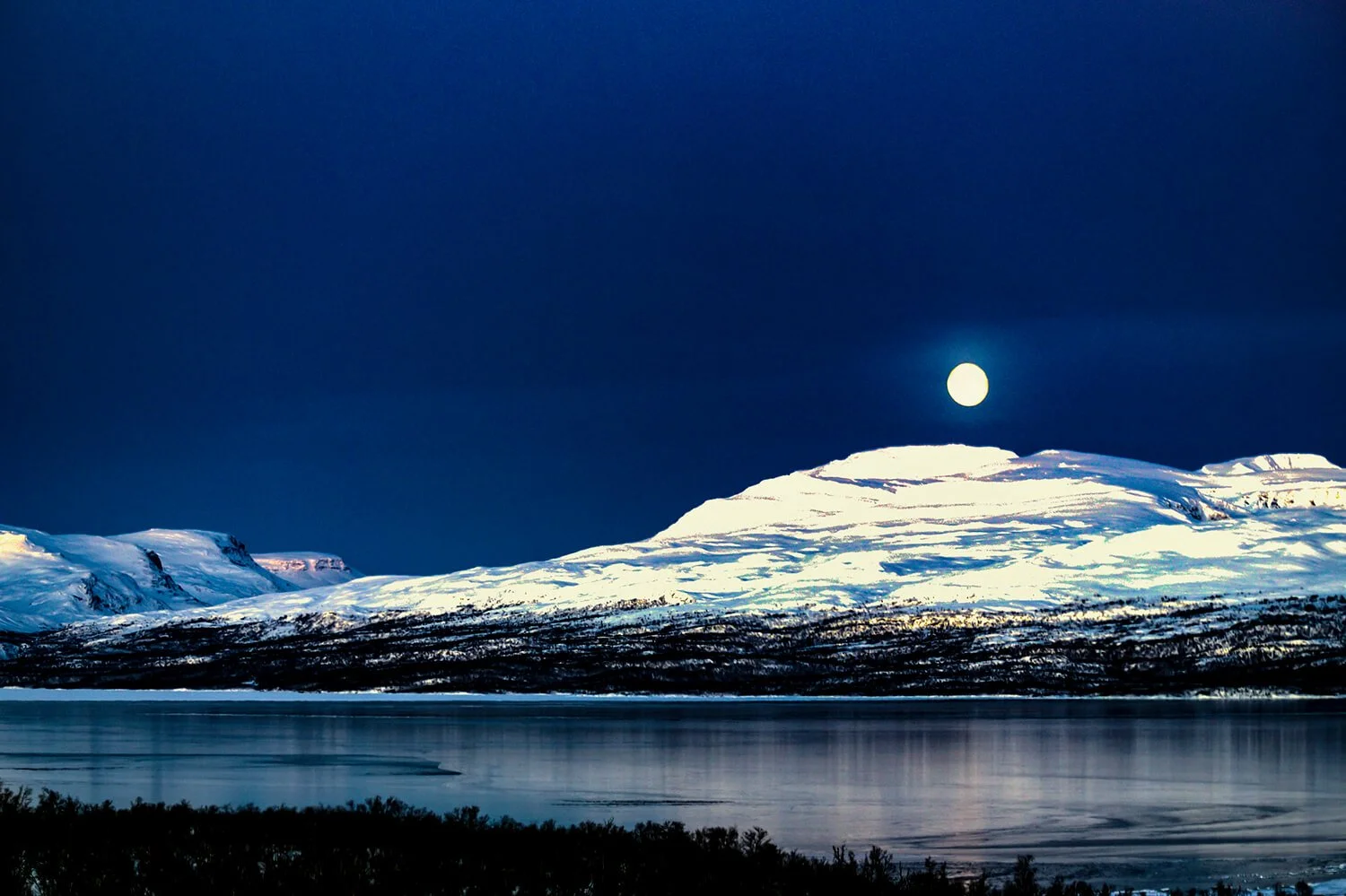 Snow-covered mountains under a dark night sky illuminated by a bright full moon,.  This photo was taken in Abisko, Sweden. Abisko is located inside the Arctic Circle in Northern Sweden.