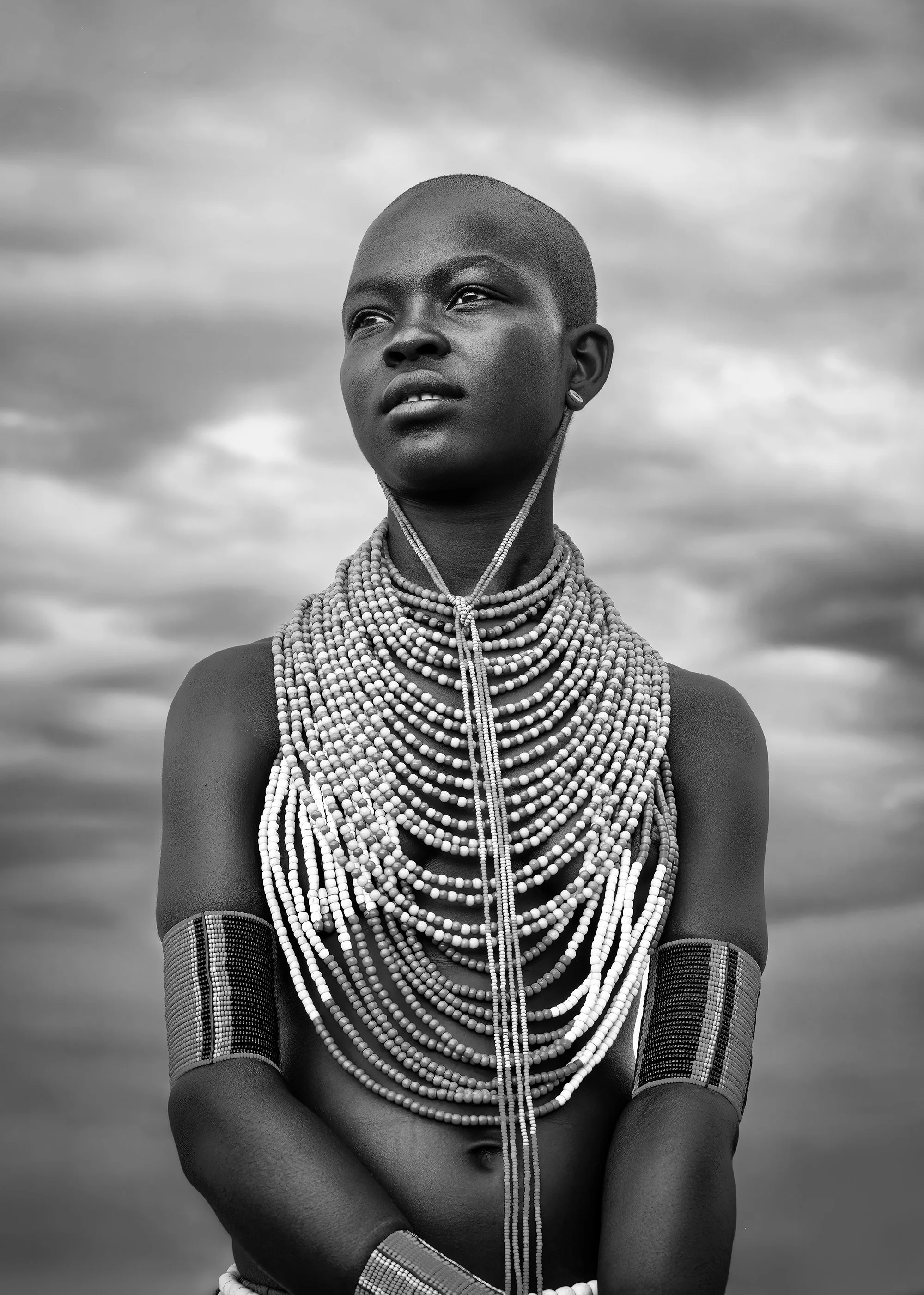 A young woman from an African tribe wearing multiple beaded necklaces and arm cuffs, standing outdoors with a cloudy sky in the background.