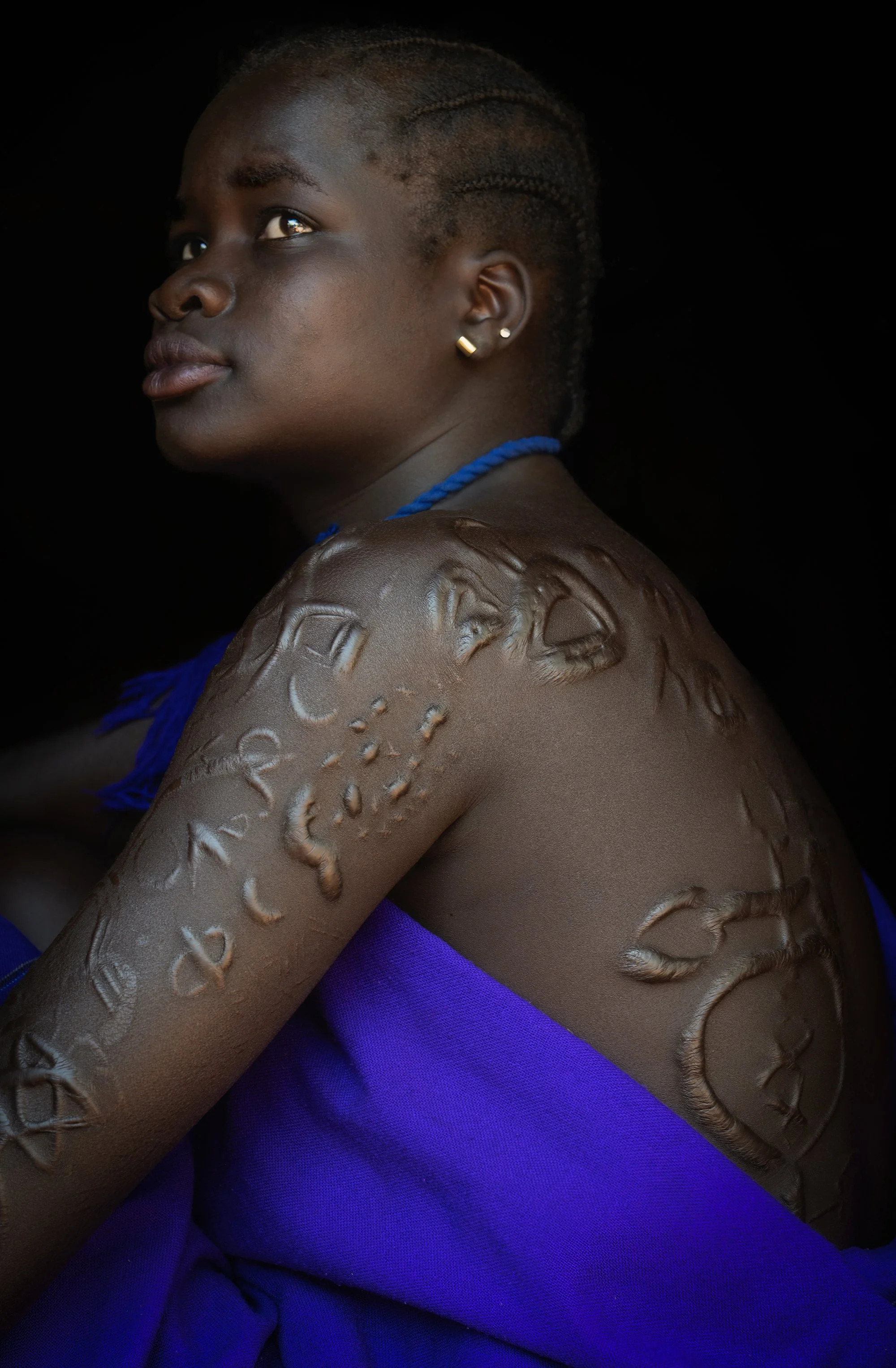 A woman with short, braided hair and earrings, wearing a purple cloth, has tribal-style body scars or markings on her shoulder and upper arm, set against a black background.