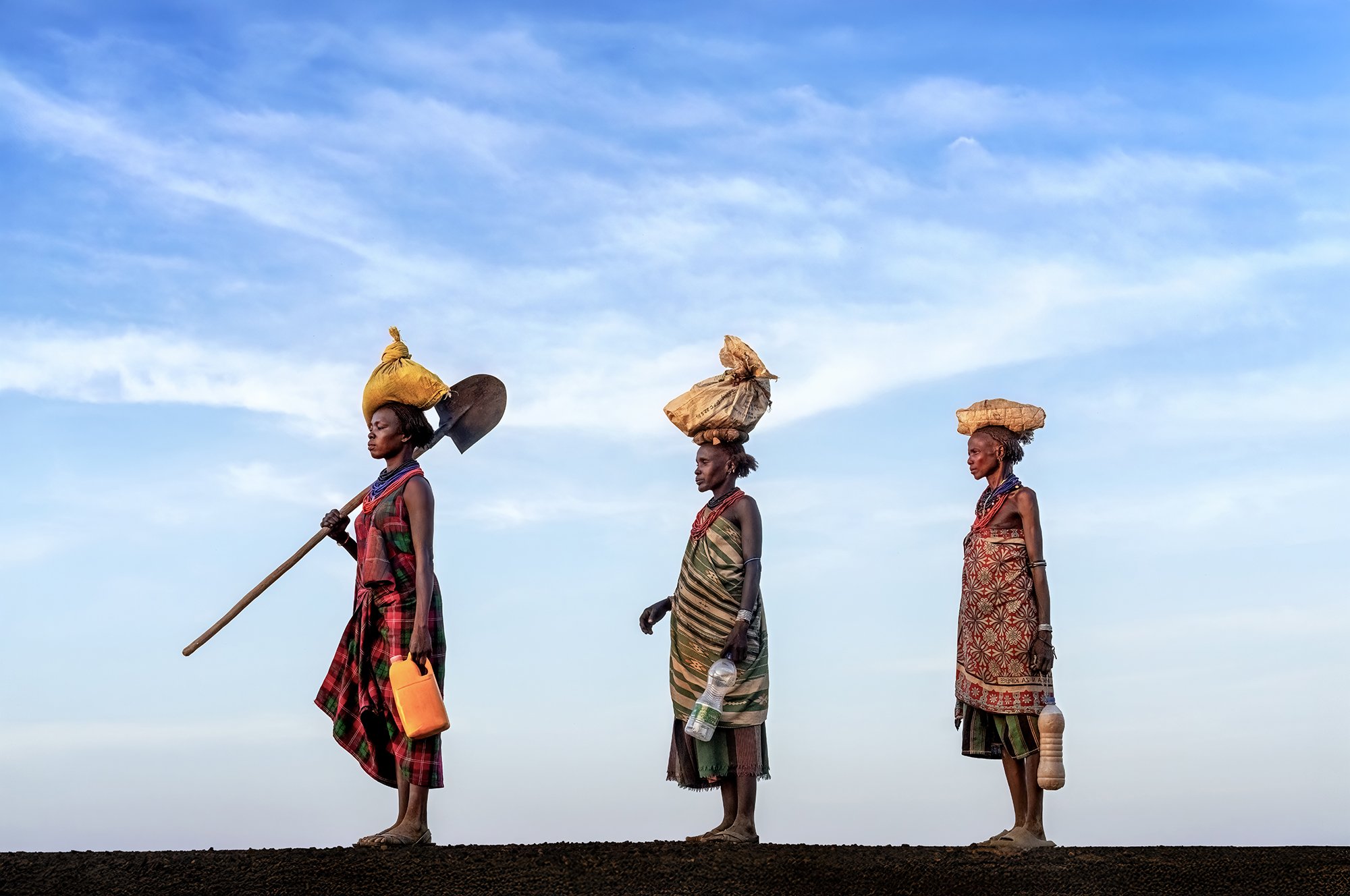 Three women in traditional clothing walking on a dirt path, carrying bags on their heads, with a blue sky and clouds in the background.