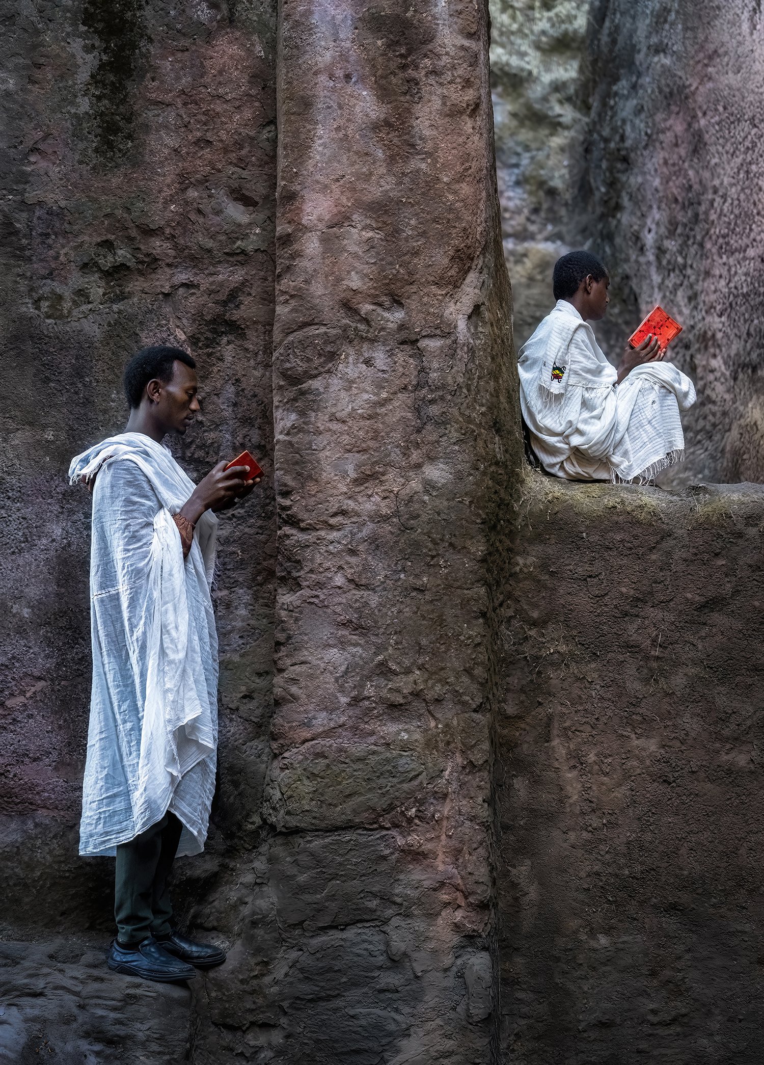 Two men dressed in white robes sitting and standing on rocky ledges, each holding a small red book, in front of a rugged rock wall.