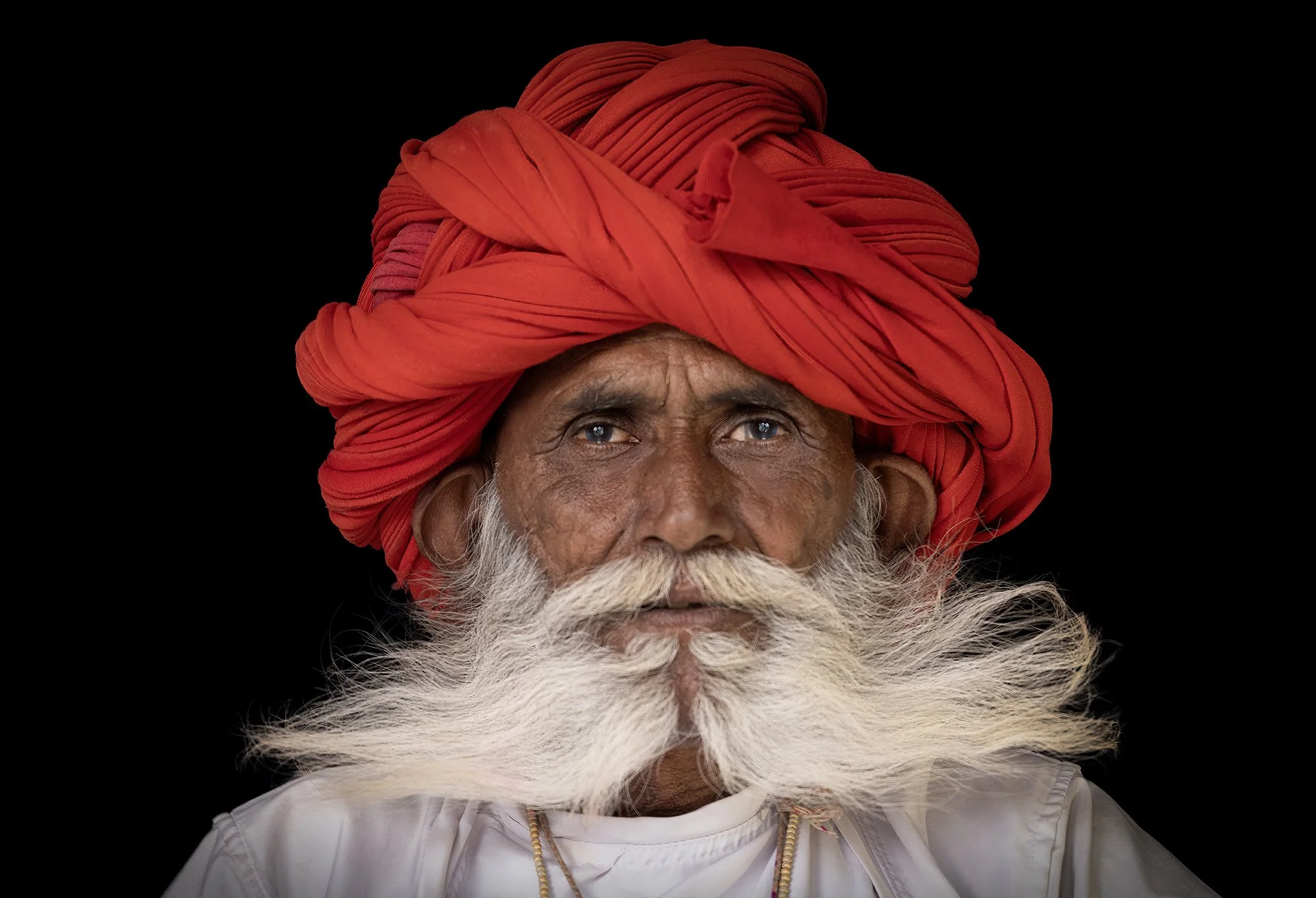 Close-up portrait of an elderly man with a long white beard, wearing a large red turban and a traditional white garment, against a black background.