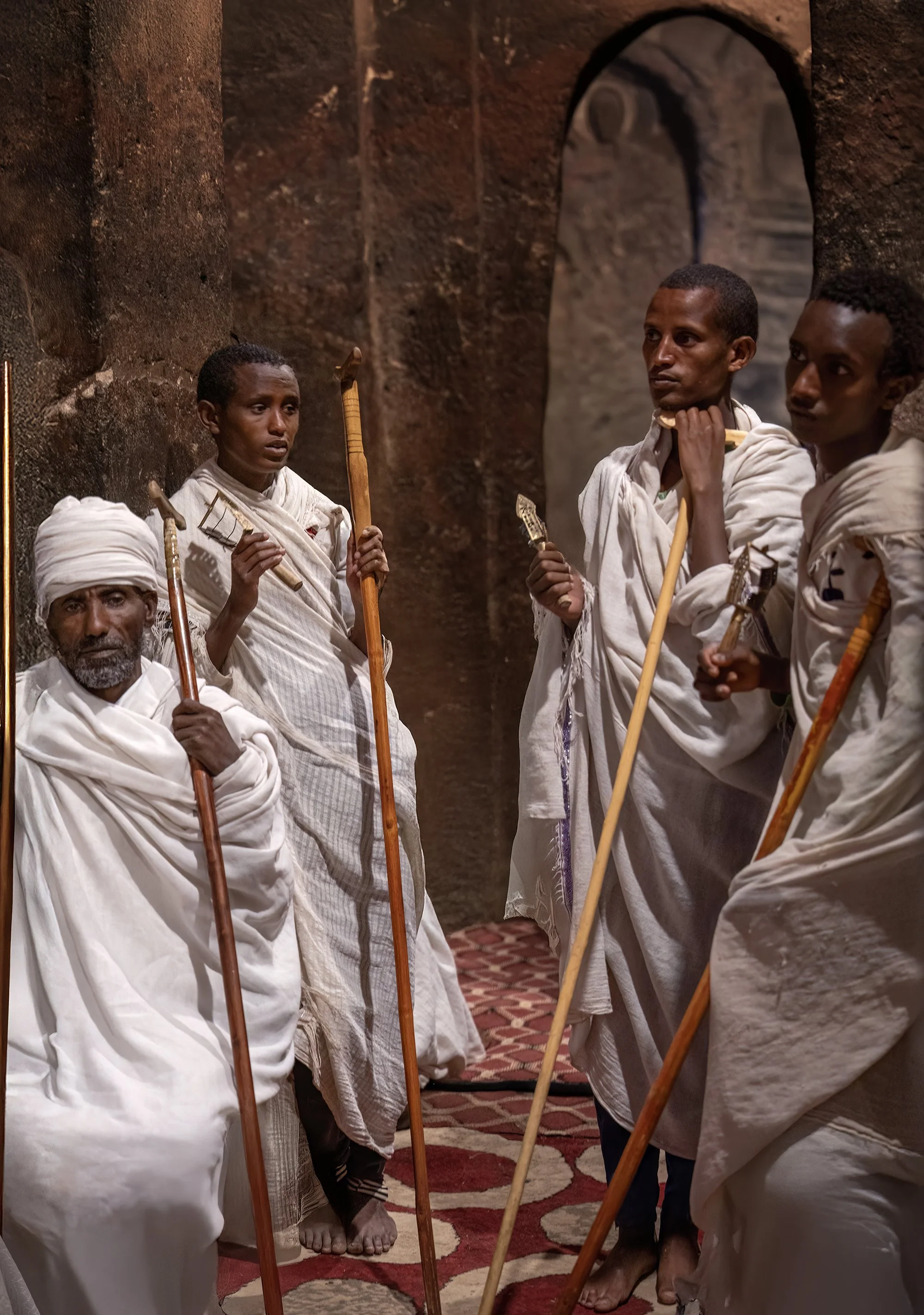 A group of five Ethiopian men dressed in traditional white clothing, standing indoors against a dark brick wall. They are holding wooden staffs and small objects in their hands, appearing to be in a serious discussion or ceremony.