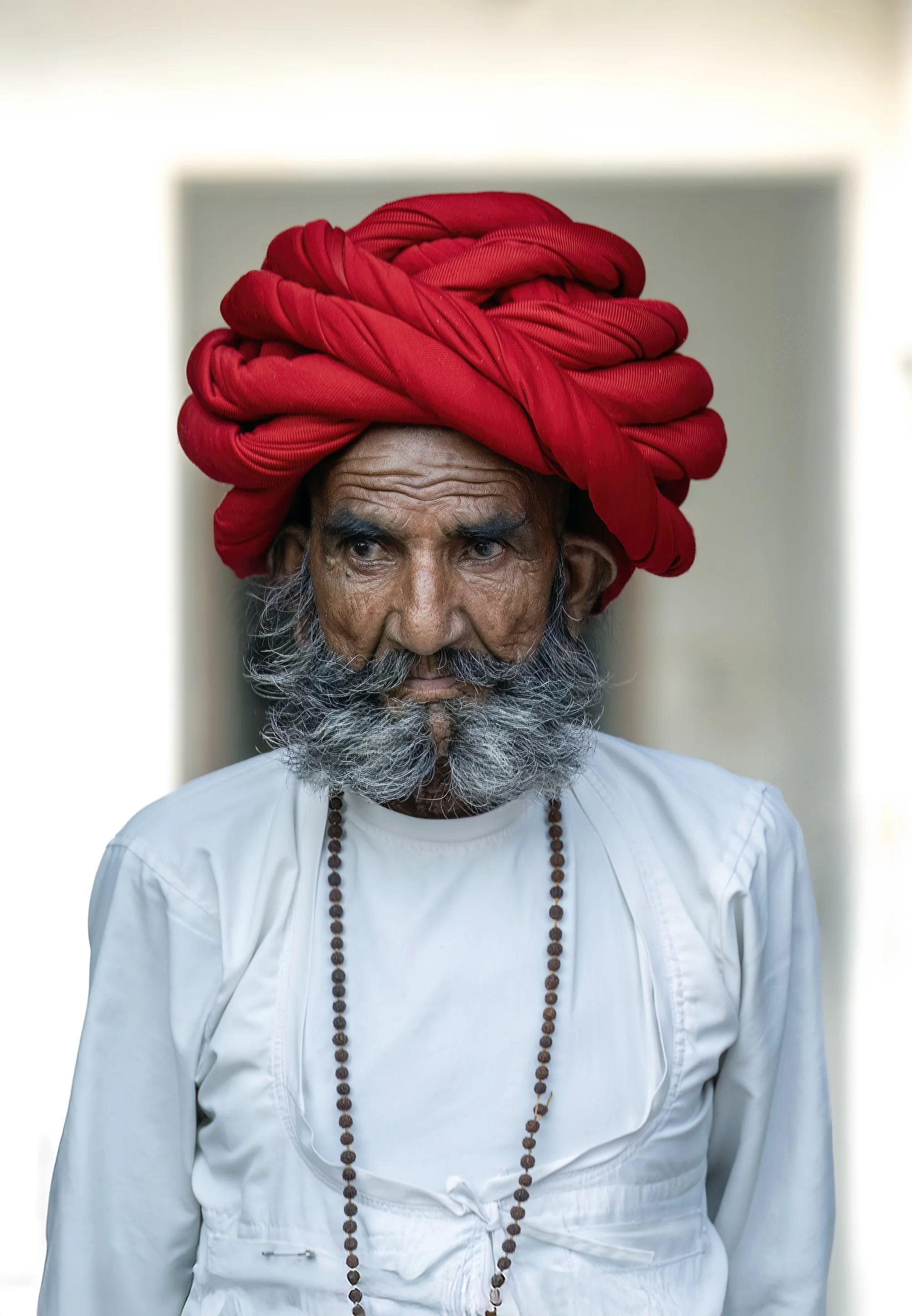 An elderly man with a white beard wearing a white traditional outfit and a long beaded necklace, with a red turban wrapped around his head.