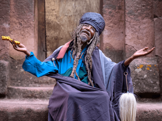 A man with long dreadlocks and a gray beanie, wearing a blue and gray robe, sitting on stone steps with both arms raised, holding a yellow guitar in one hand, with a large, blonde wig hanging from his other. The background features weathered stone wa