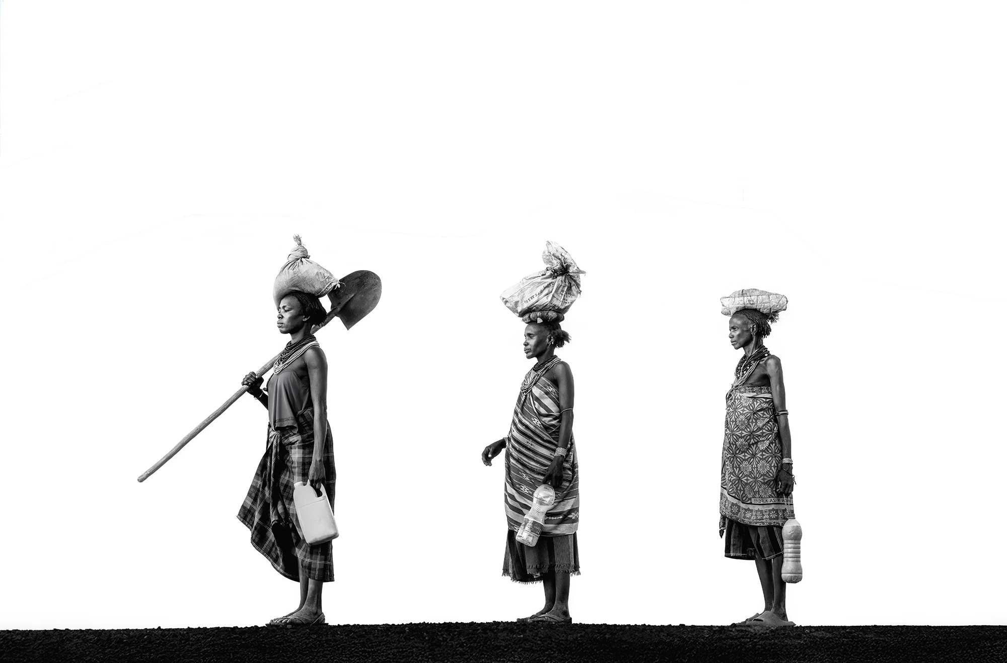 Black and white photo of three women standing on a dark surface, each carrying items on their head and holding objects in their hands. The women are dressed in traditional clothing and jewelry, with a minimalist white background.