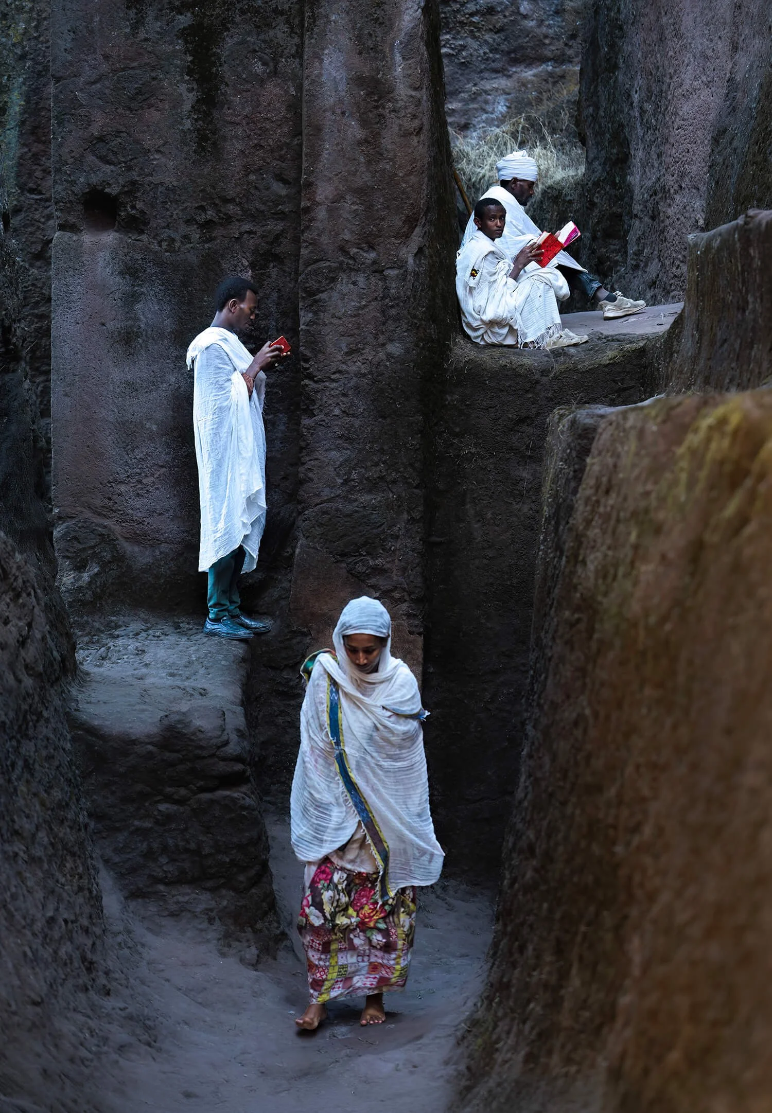 
These two devout men gather outside the sacred walls of a rock-hewn church in the heart of Lalibela, Ethiopia.  They are wearing traditional white garments.  The woman is walking to pray inside the church.  People have been praying at the Lalibela c