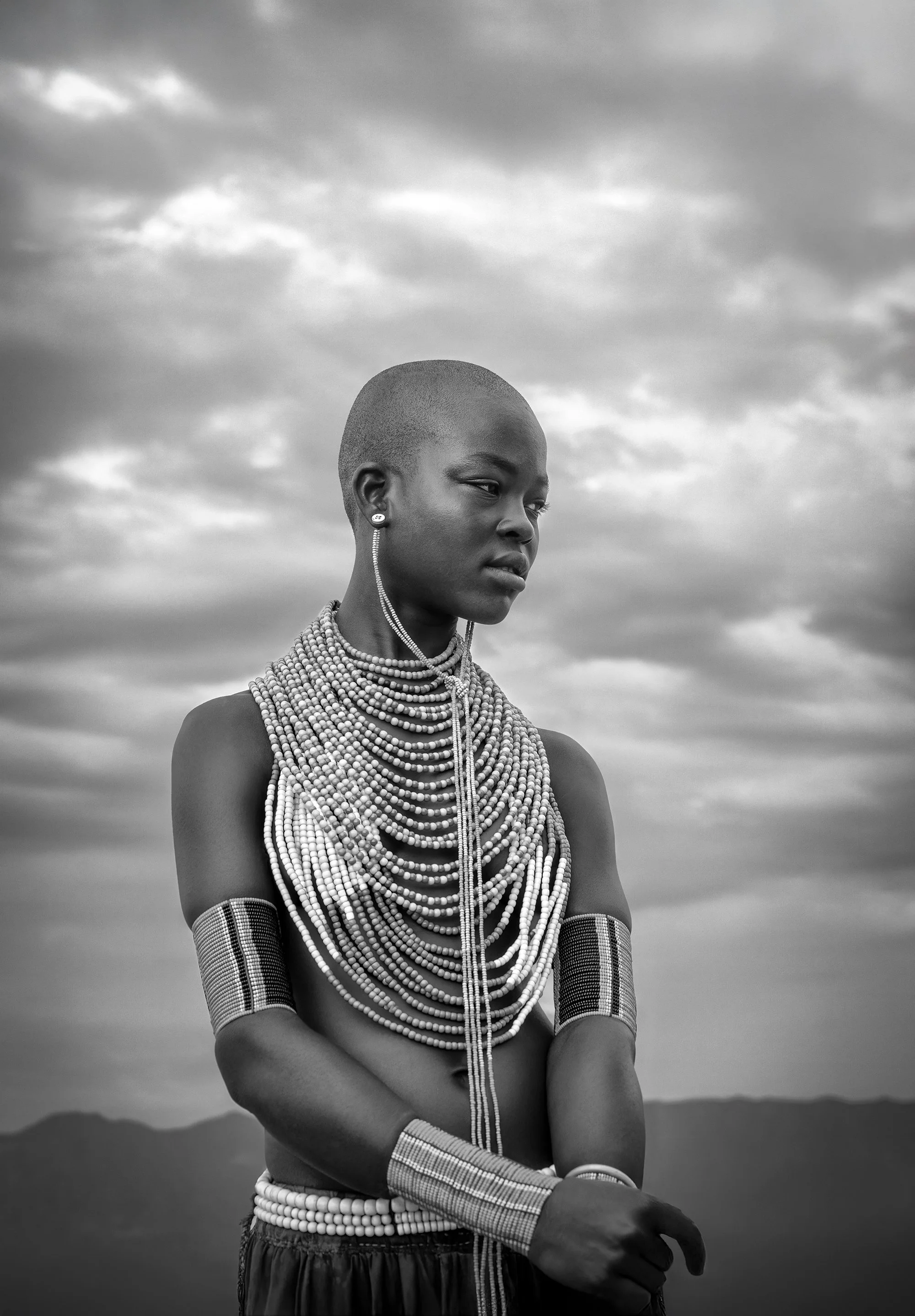 Portrait of a young woman wearing traditional jewelry, including multiple necklaces, earrings, and arm bands, standing against a cloudy sky backdrop.