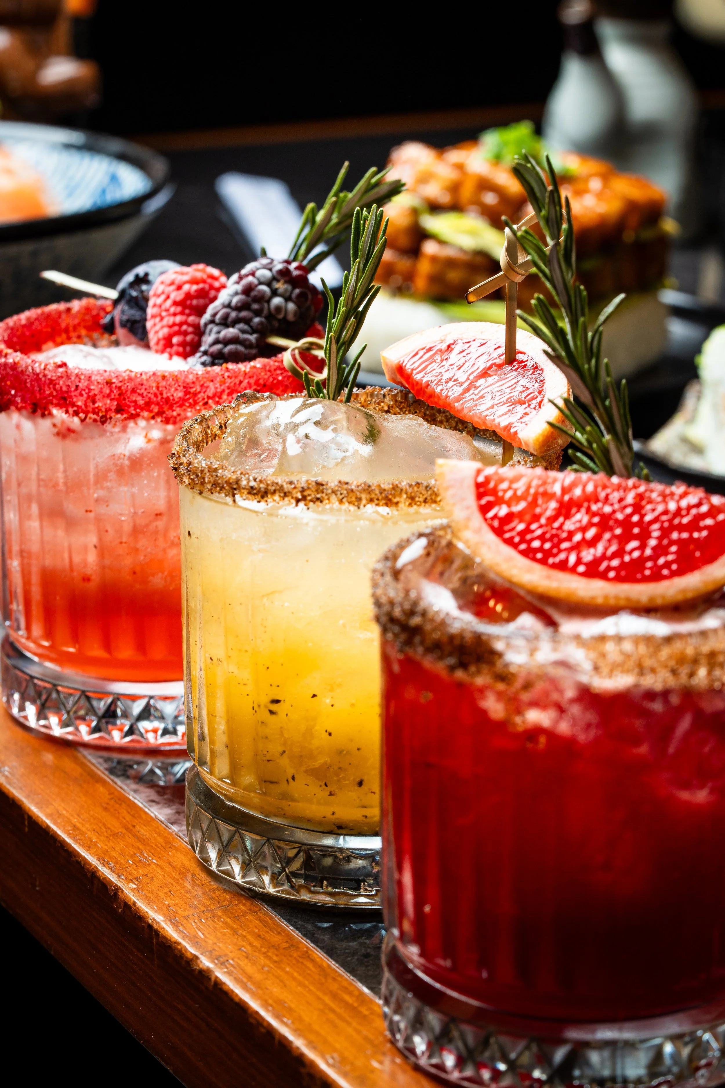 Tres cócteles con adornos de fruta y hierbas, con un fondo de comida en un restaurante.