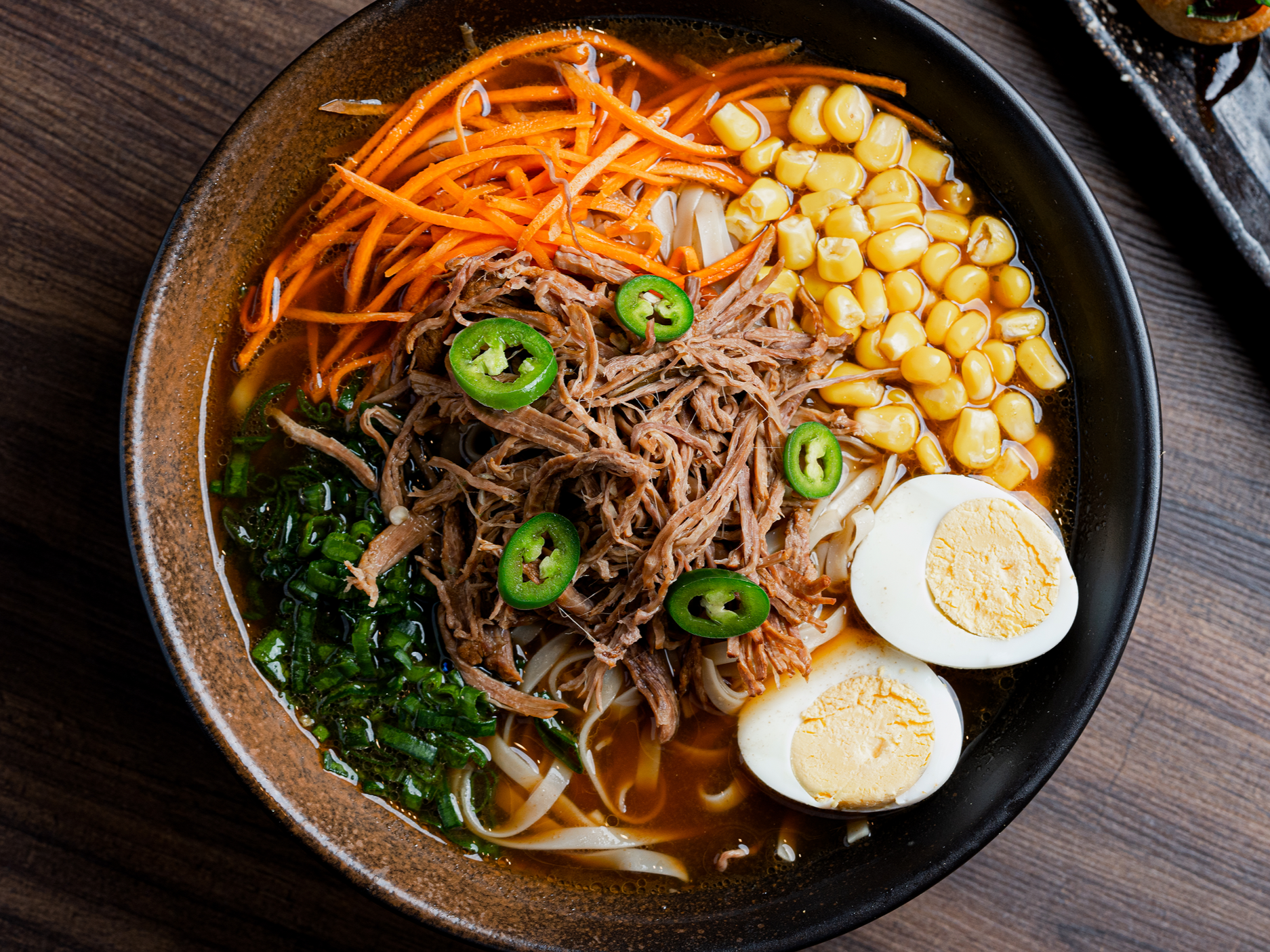 Tazón de sopa de ramen con carne desmenuzada, cebollines, jalapeños, zanahorias ralladas, maíz, huevos cocidos y fideos en caldo.