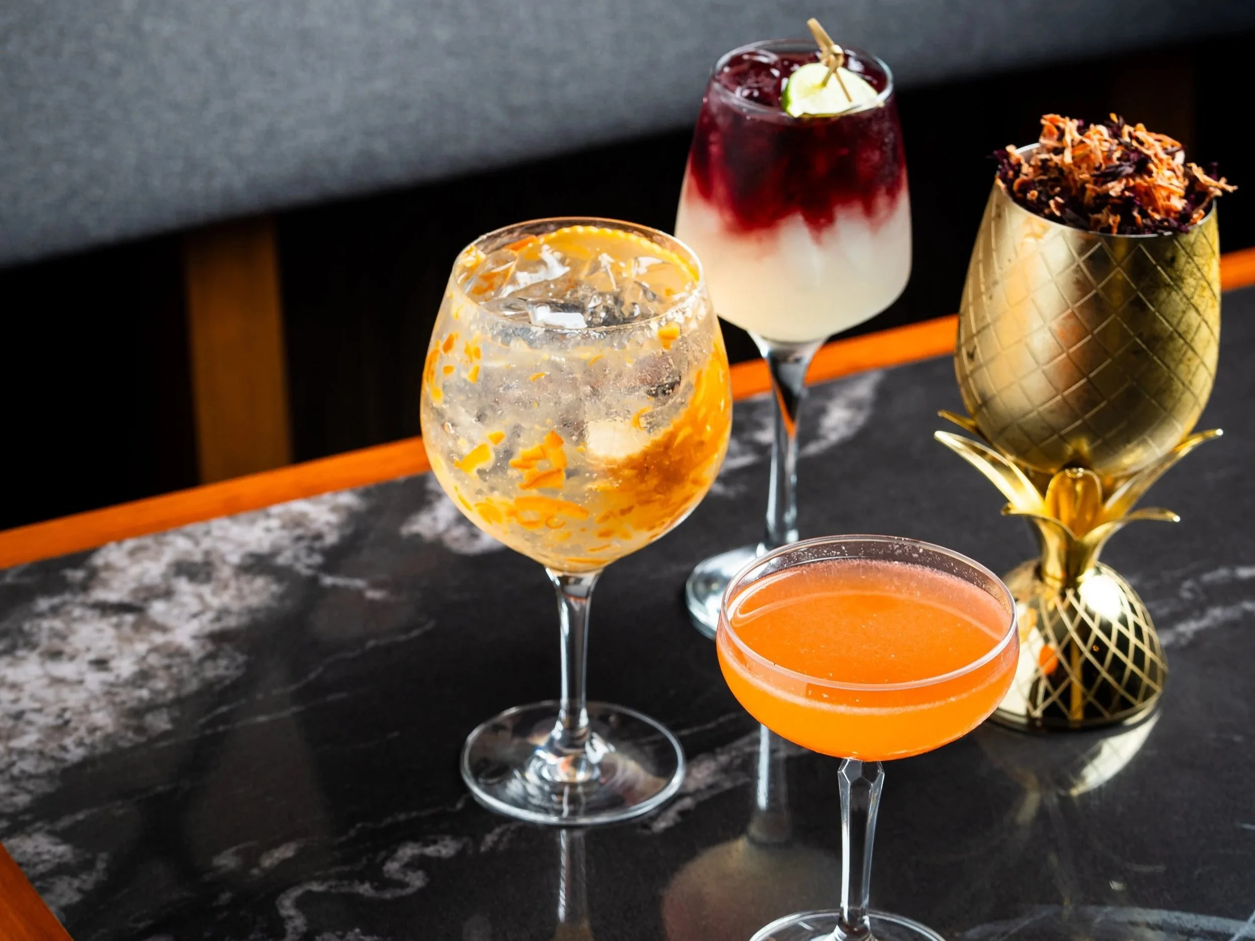 Cuatro cócteles en copas elegantes con diferentes colores y decoraciones, junto a una piña dorada decorativa en una mesa de mármol oscuro.