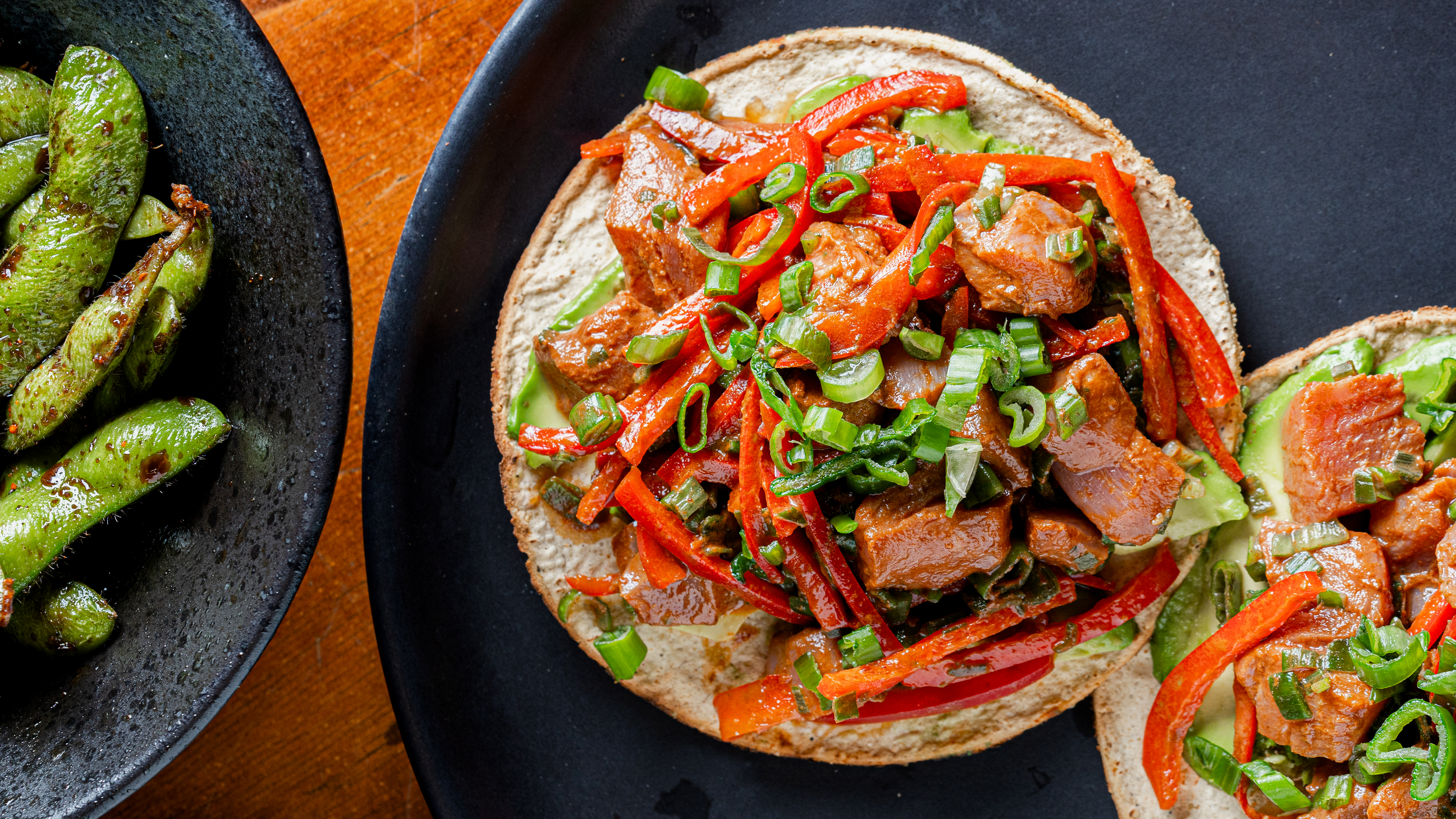Tostada con carne y verduras, acompañada de edamames en plato negro.