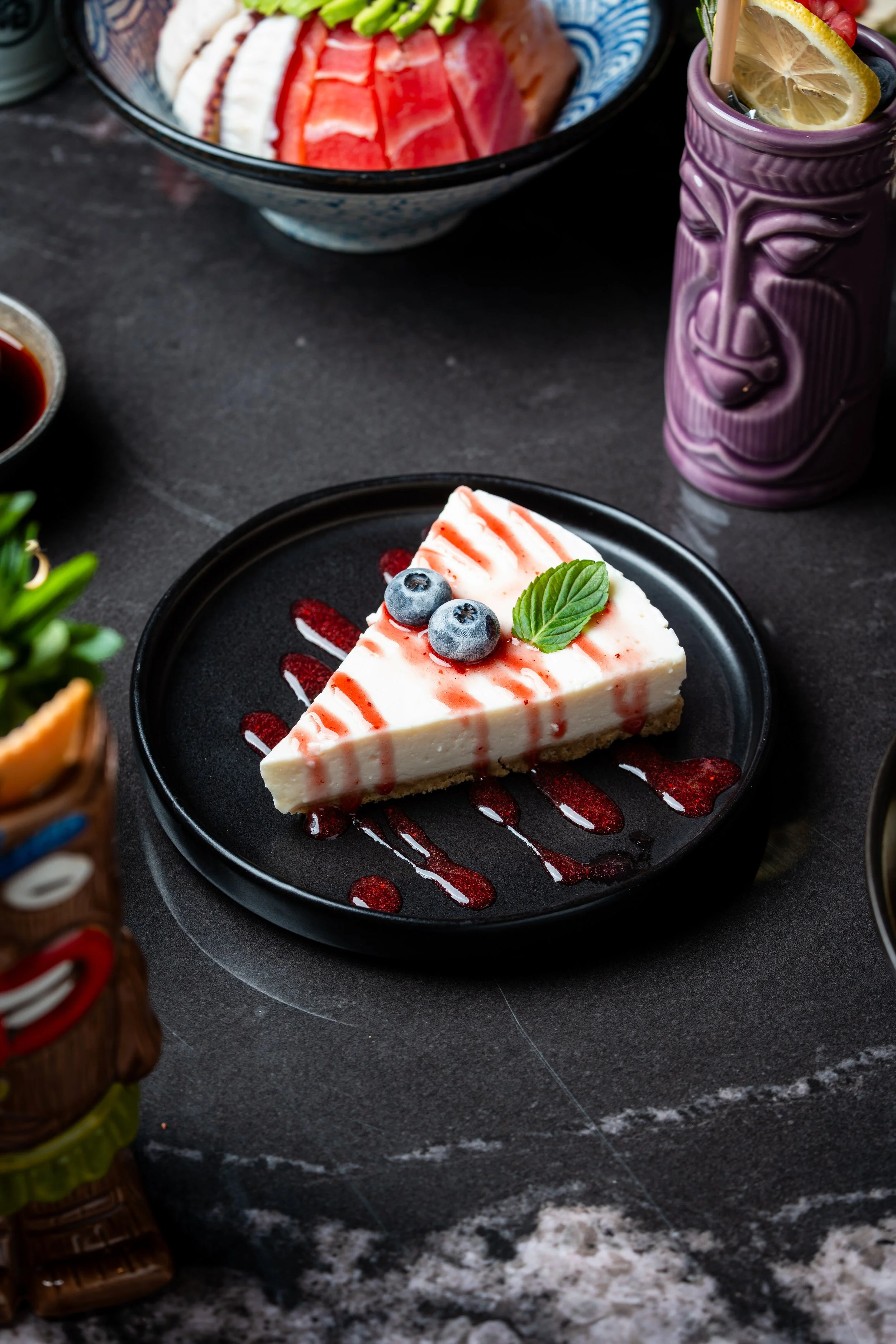 Porción de pastel de queso decorada con arándanos, menta y salsa de frutos rojos en un plato negro.