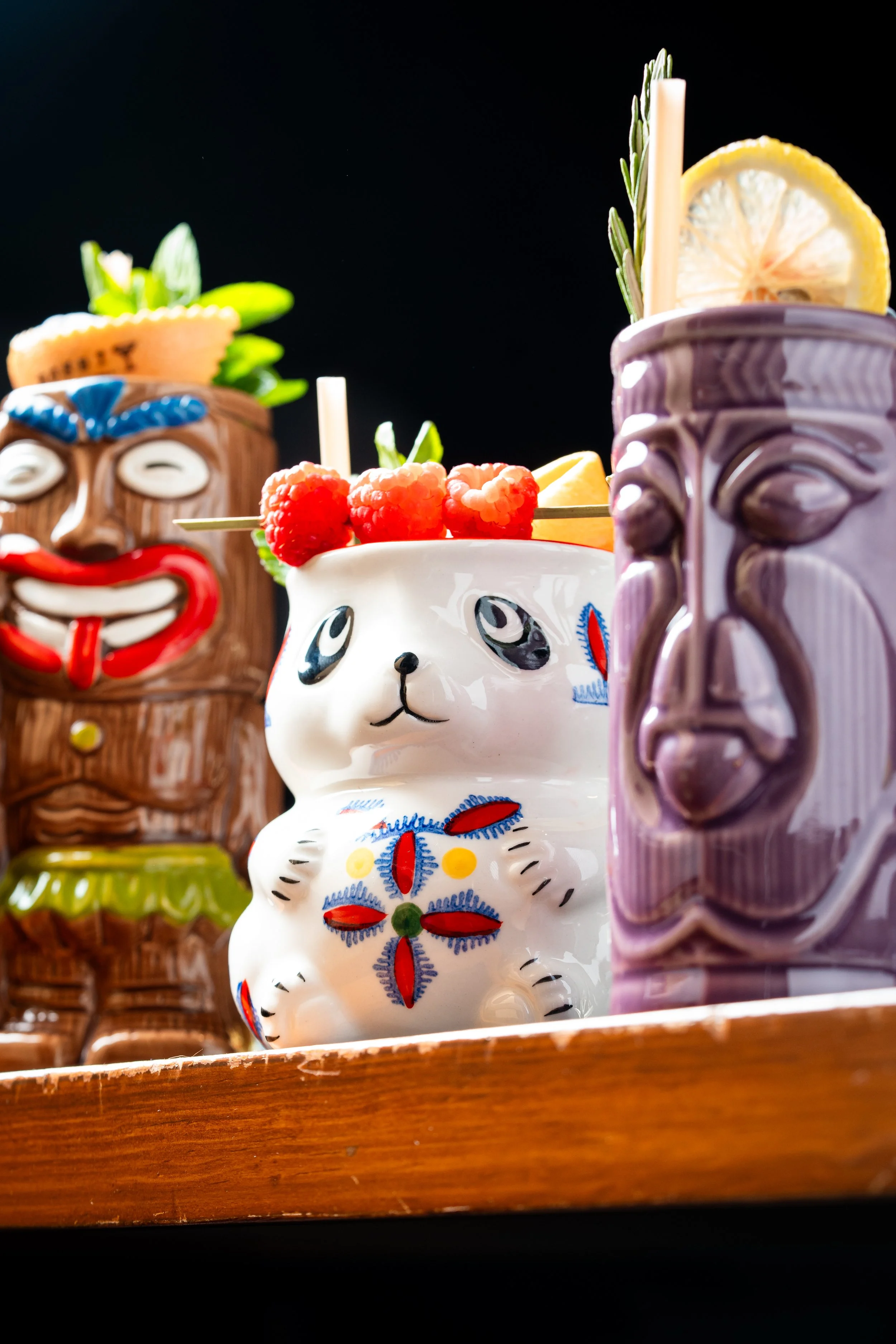 Tres vasos con decoración de estilo tiki en forma de caras, con fruta y pajillas, sobre una superficie de madera.