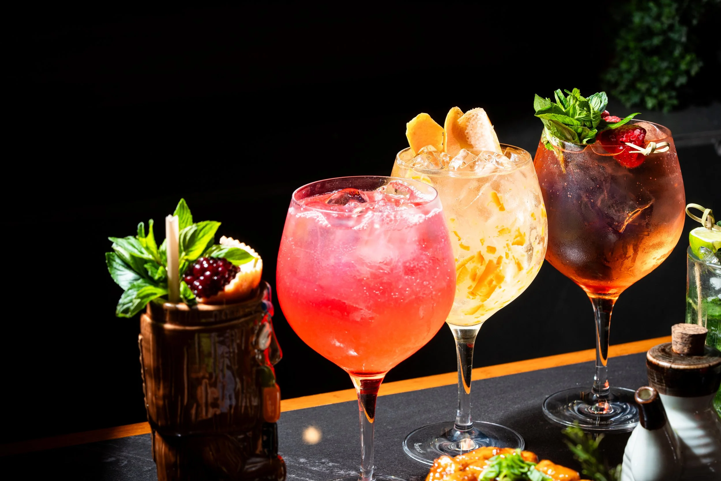 Tres copas de cóctel con hielo y decoraciones de fruta y hierbas, en un fondo oscuro.