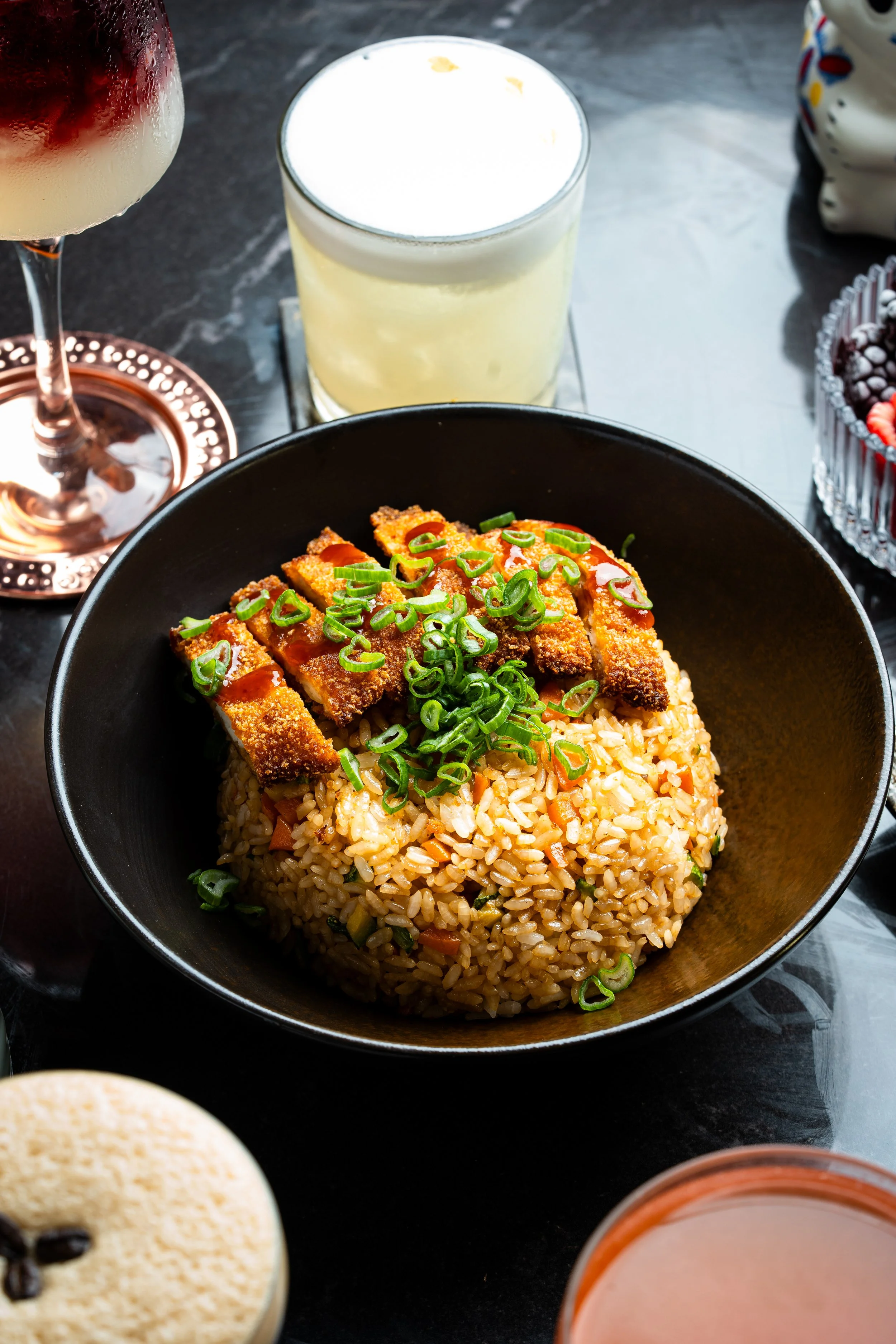 Un tazón con arroz frito con vegetales, cubos de pollo empanizado y cebollines cortados en rodajas, acompañado de bebidas y postres en una mesa.
