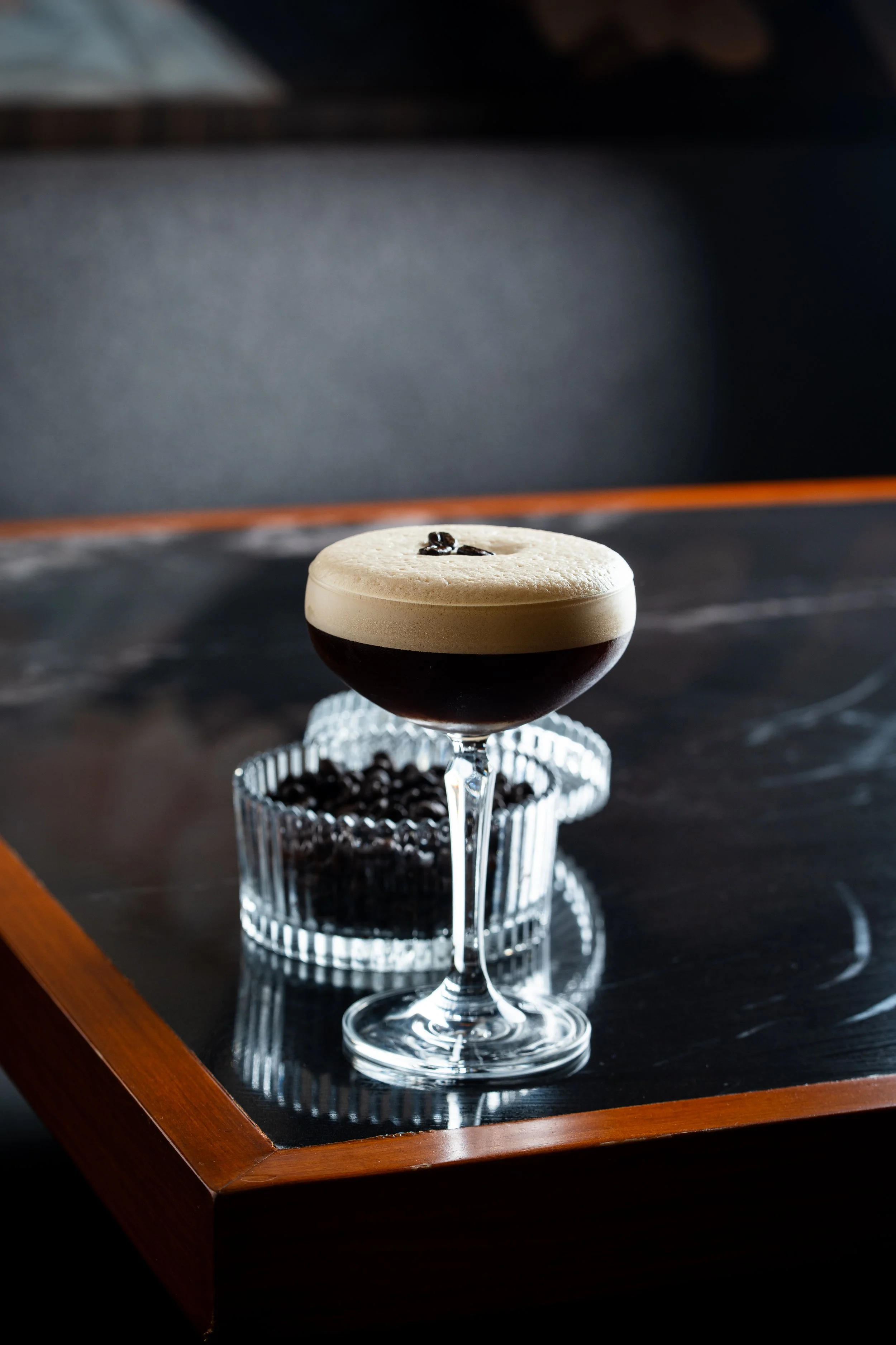 Cóctel oscuro con espuma cremosa y granos de café en la parte superior, servido en una copa de cóctel con un fondo de recipiente con granos de café.