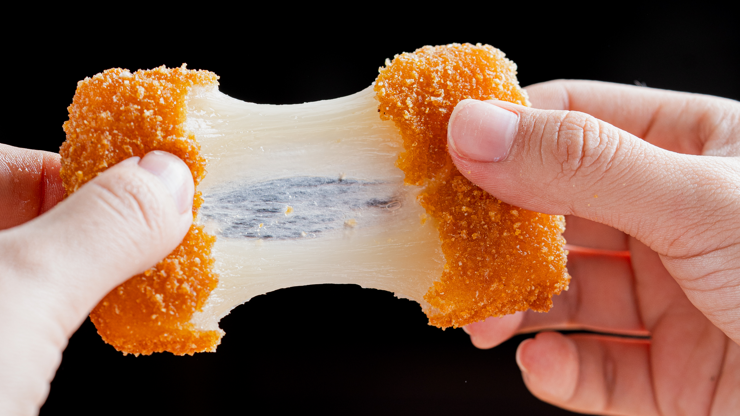 Manos sosteniendo una pieza de pan frito con queso derretido en el interior, sobre fondo negro.