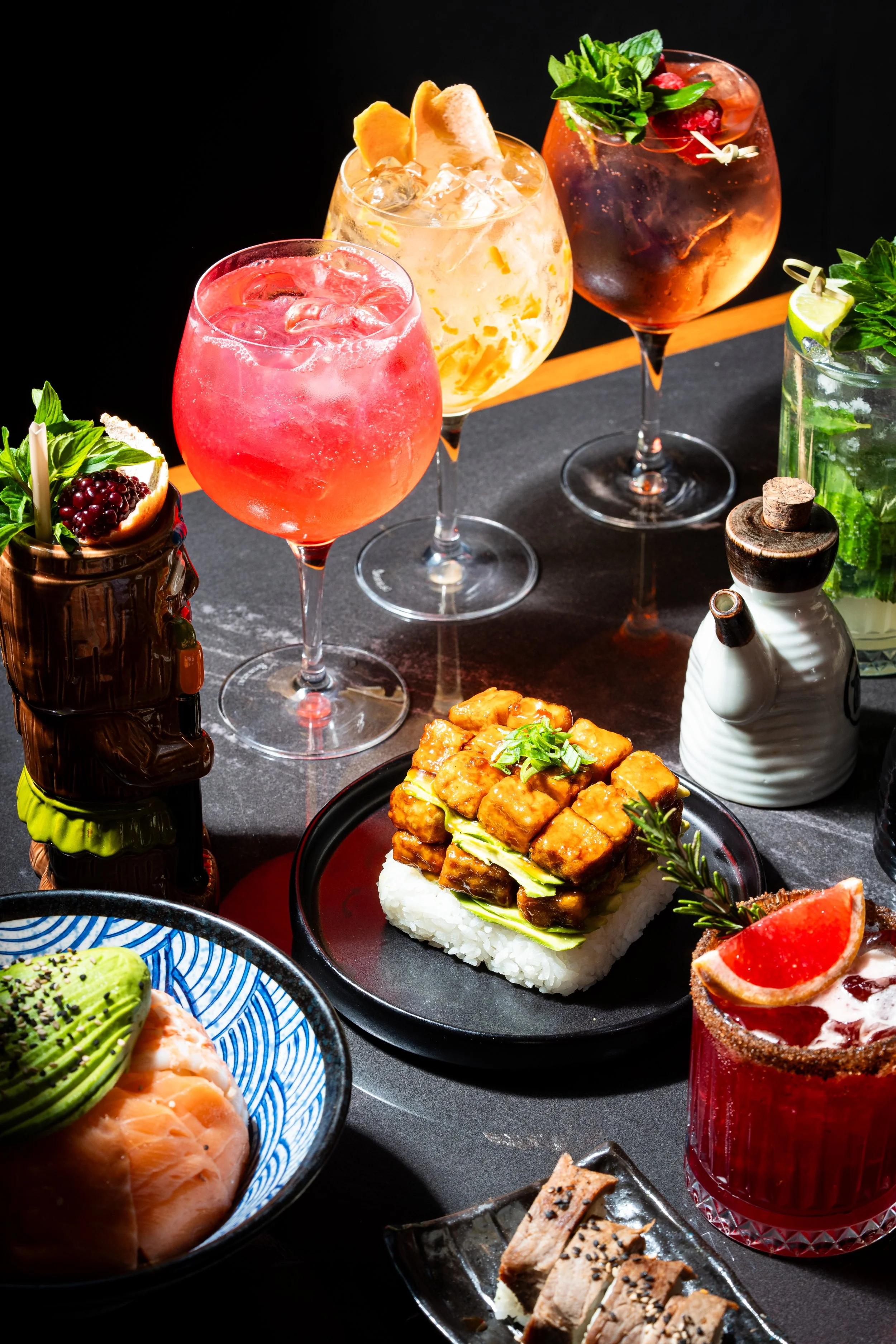 Una variedad de cócteles coloridos y comida japonesa en una mesa oscura.