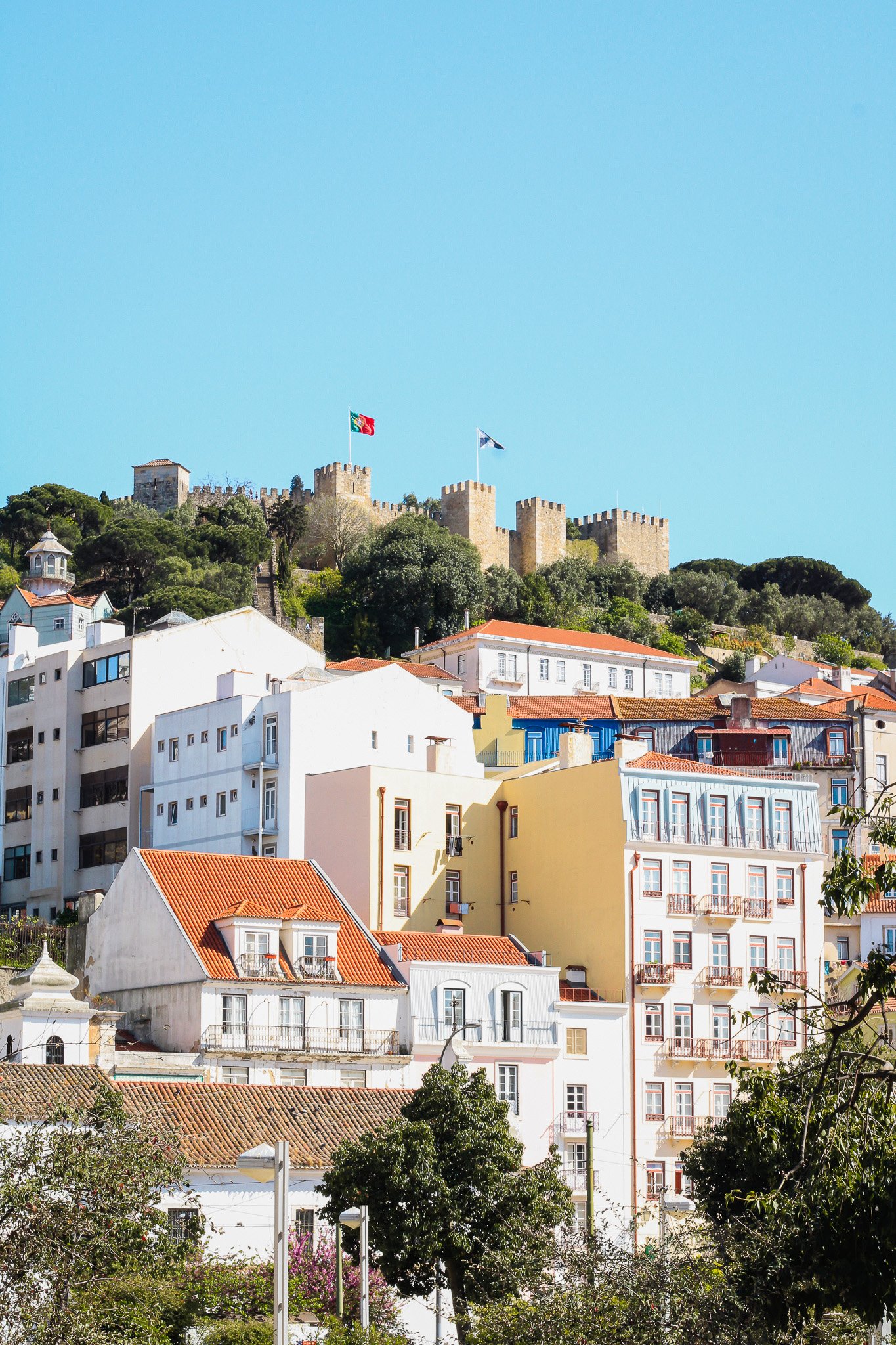 Casas coloridas y un castillo en una colina con banderas ondeando, árboles verdes alrededor.