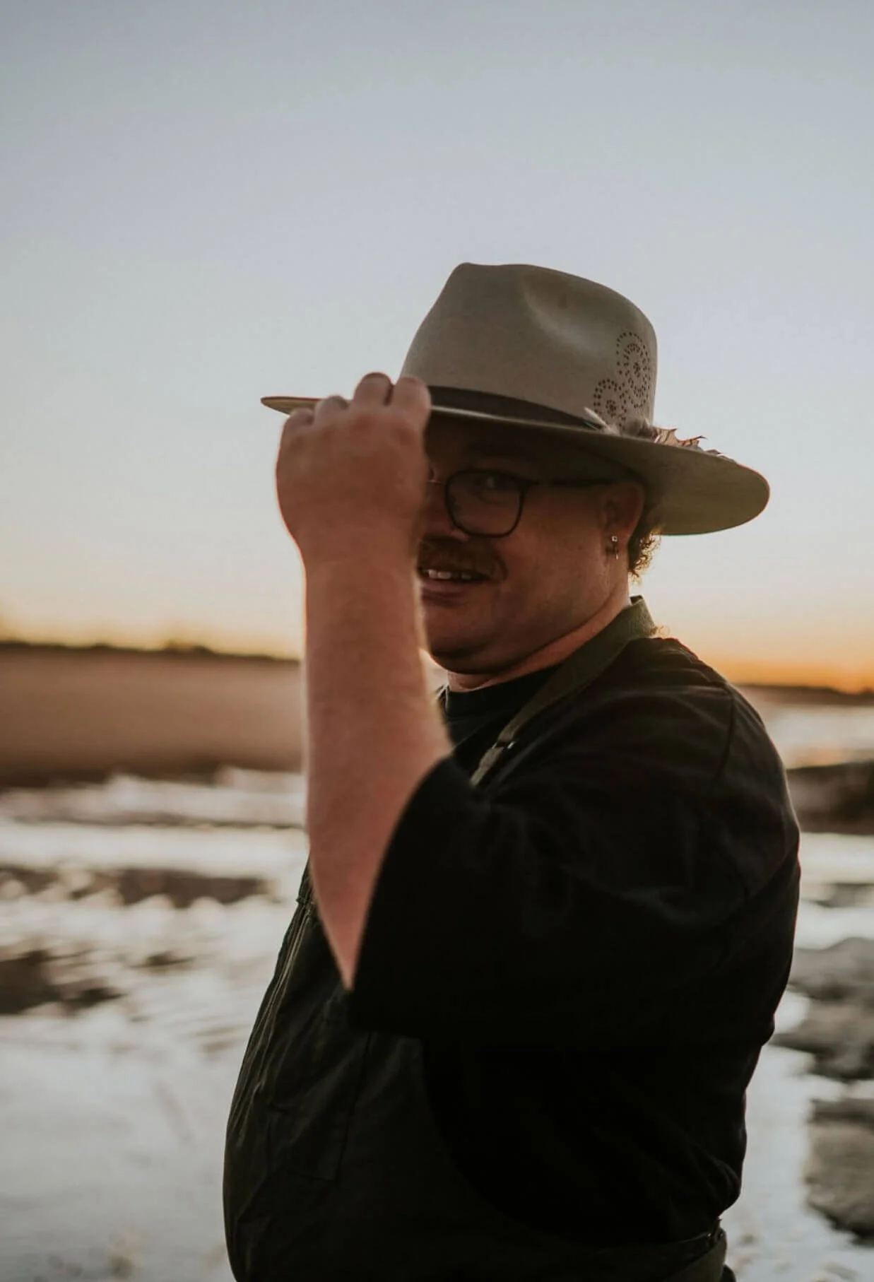 A man with glasses and a mustache wearing a wide-brimmed hat and dark clothing, lifting the hat in a gesture, standing near water during sunset.
