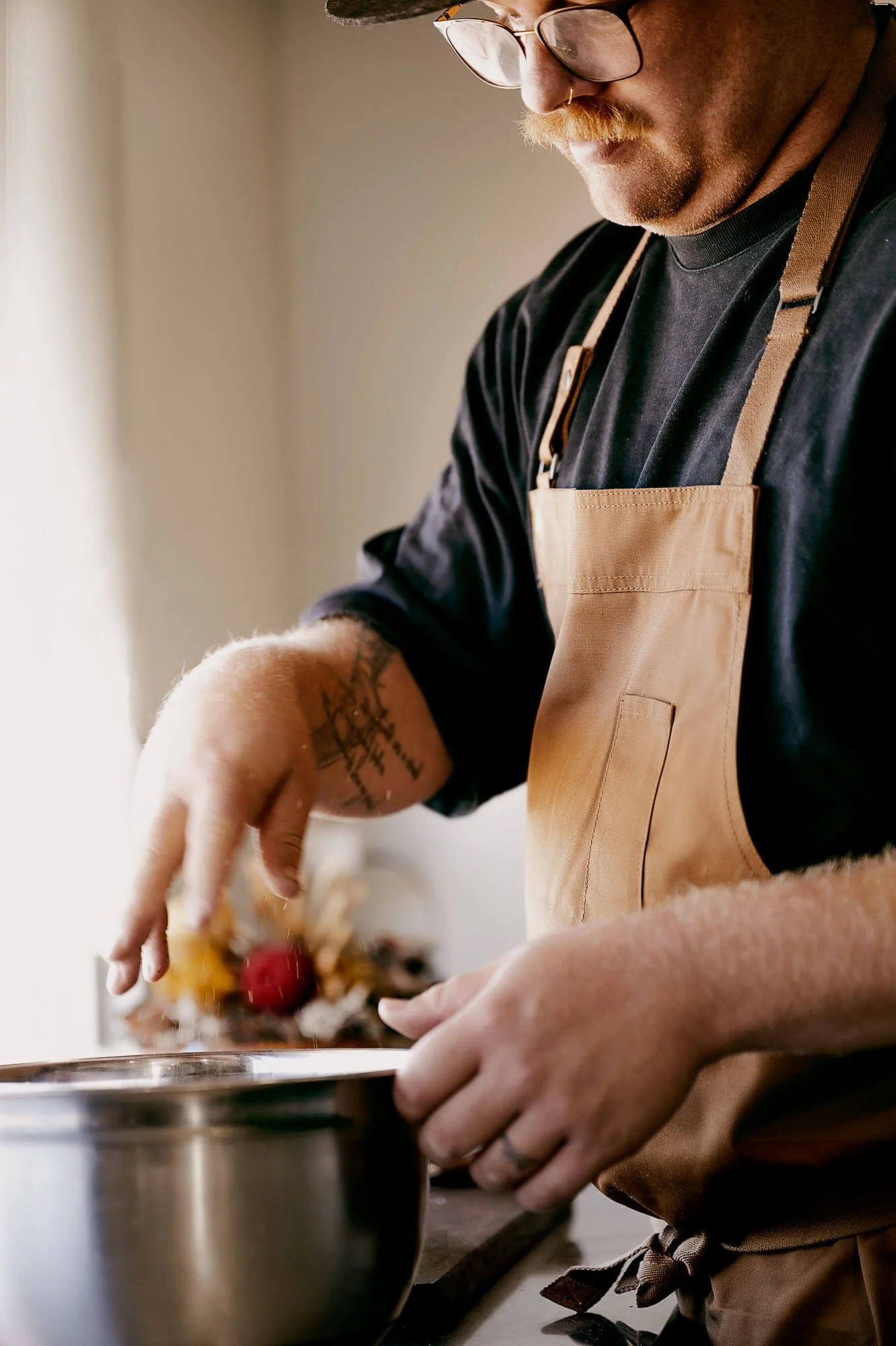A person wearing glasses, a black shirt, and a tan apron prepares food in a kitchen, with focus on their hands and torso.