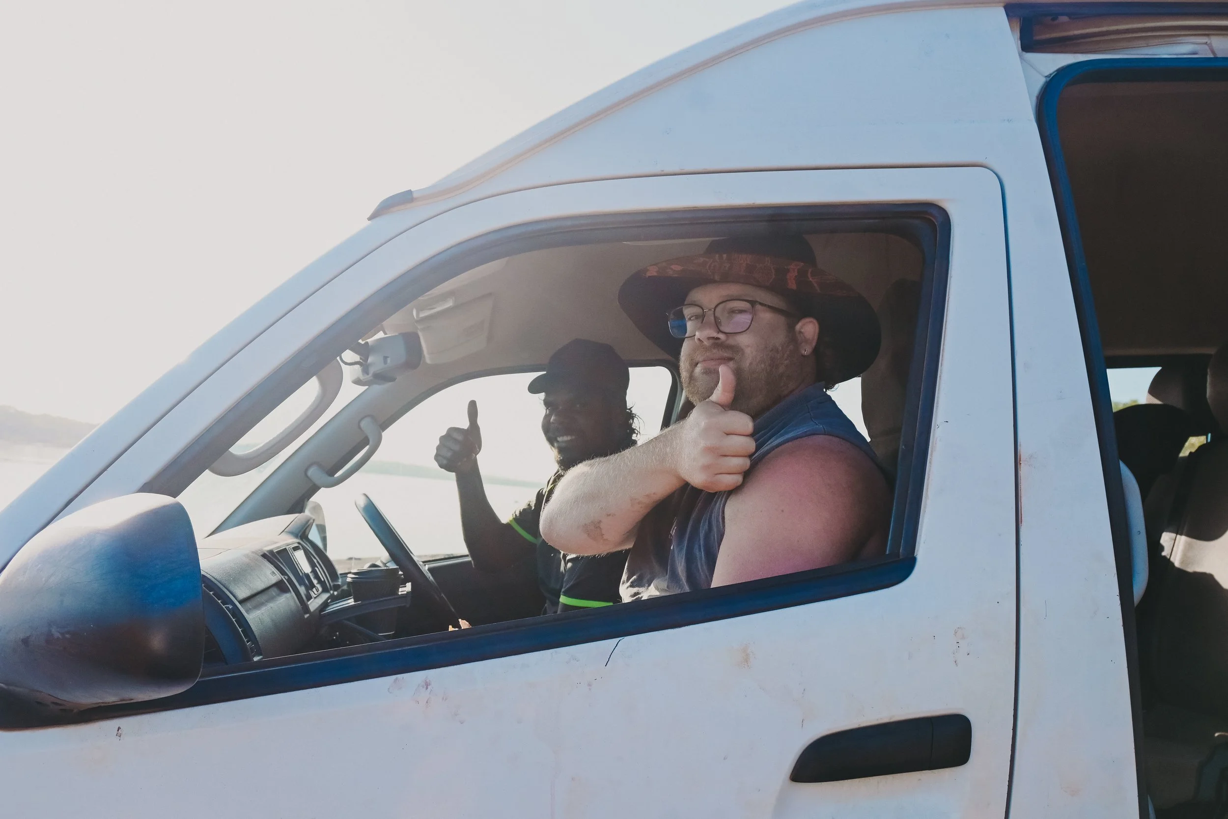 Two men sitting in a white vehicle, one wearing a straw hat and giving a thumbs-up, the other wearing a baseball cap and making a thumb-up gesture, both smiling.