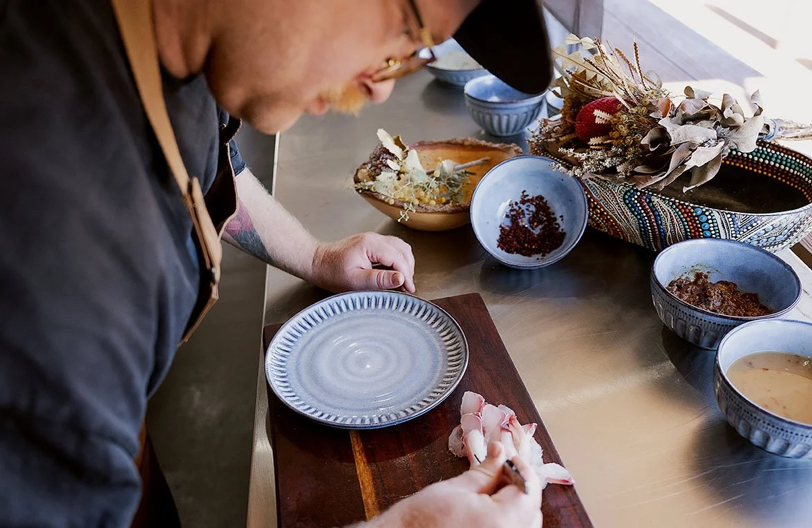 A person wearing glasses and a cap prepares to place sliced raw meat on a plate at a kitchen counter, with various bowls and a bowl of dried herbs or spices nearby.