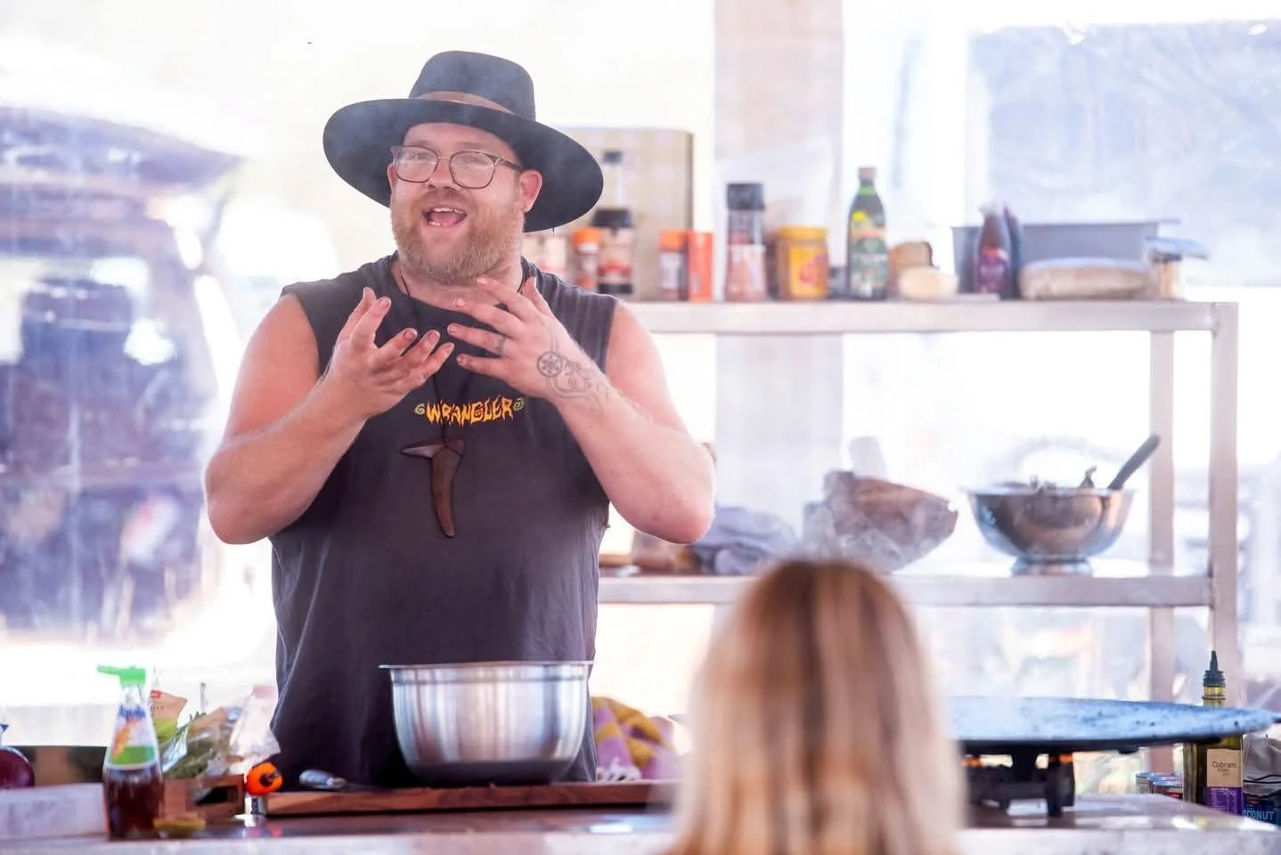 Man with glasses, beard, and tattoos wearing a wide-brimmed hat and sleeveless shirt, speaking animatedly in a kitchen or workshop setting.