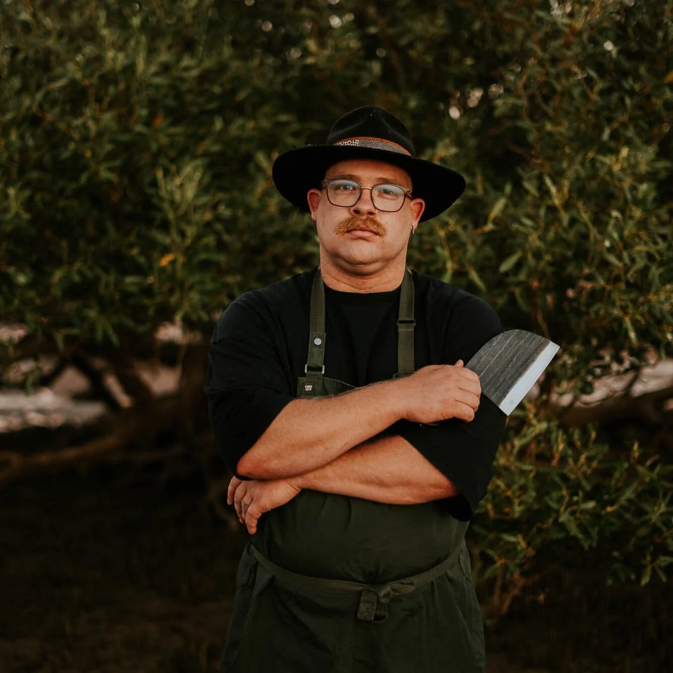 A man with glasses, a mustache, and earring standing outdoors in front of greenery, wearing a wide-brimmed hat and apron, holding a meat cleaver across his chest.