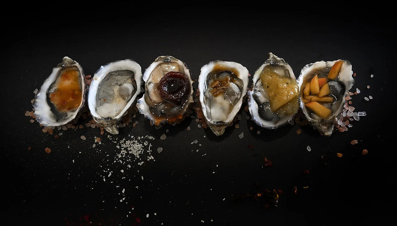Six oysters on the half shell with varied toppings, arranged in a line on a black surface, sprinkled with coarse salt and pepper.
