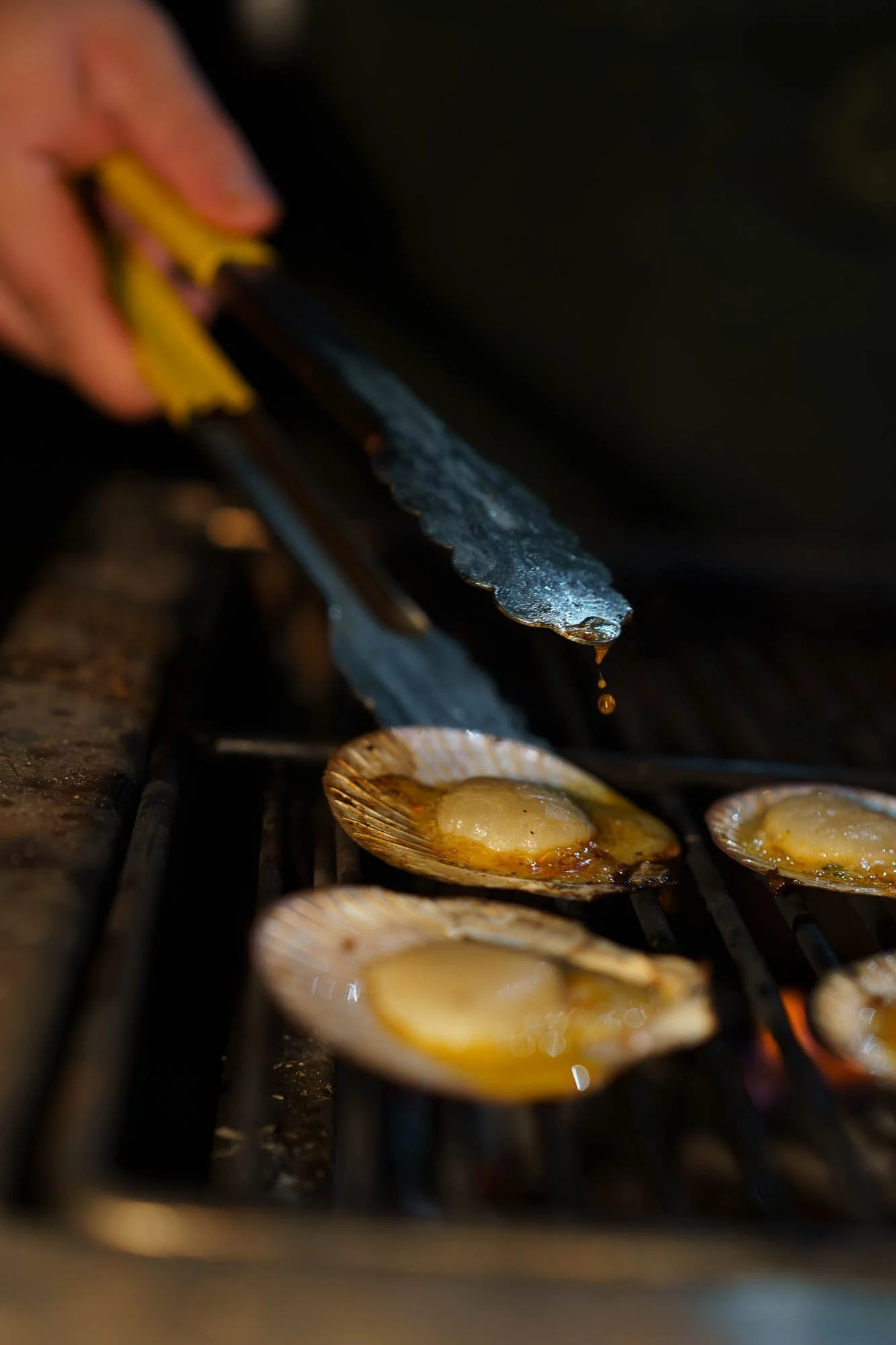 Close-up of scallops being grilled on an open flame, with a hand holding tongs in the background.