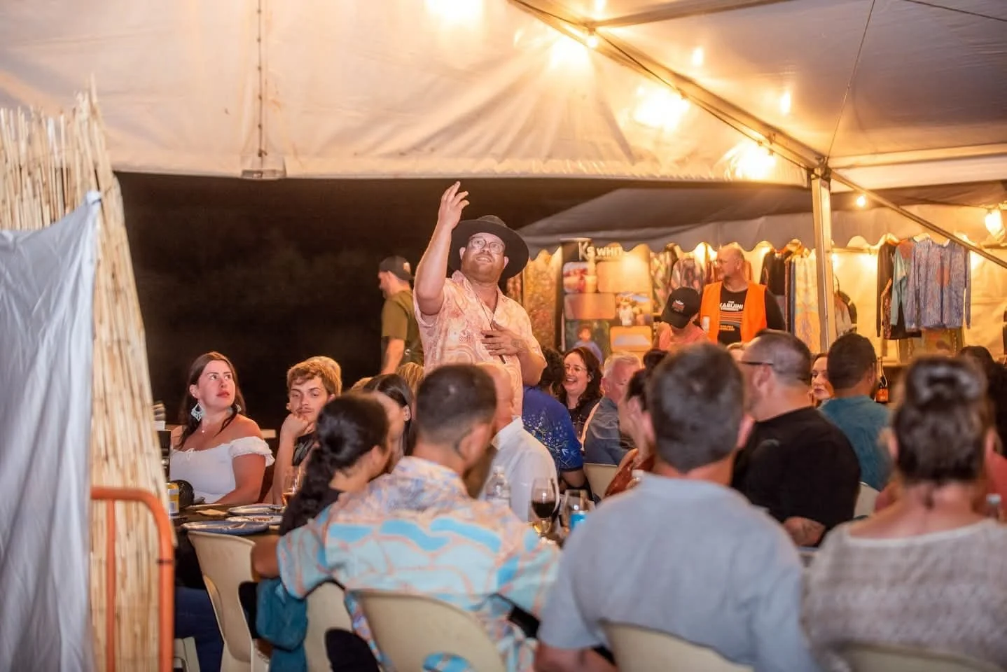 Man in a Hawaiian shirt and wide-brimmed hat speaking or singing to a seated crowd under a large tent at night, with hanging lights and colorful clothing displays in the background.