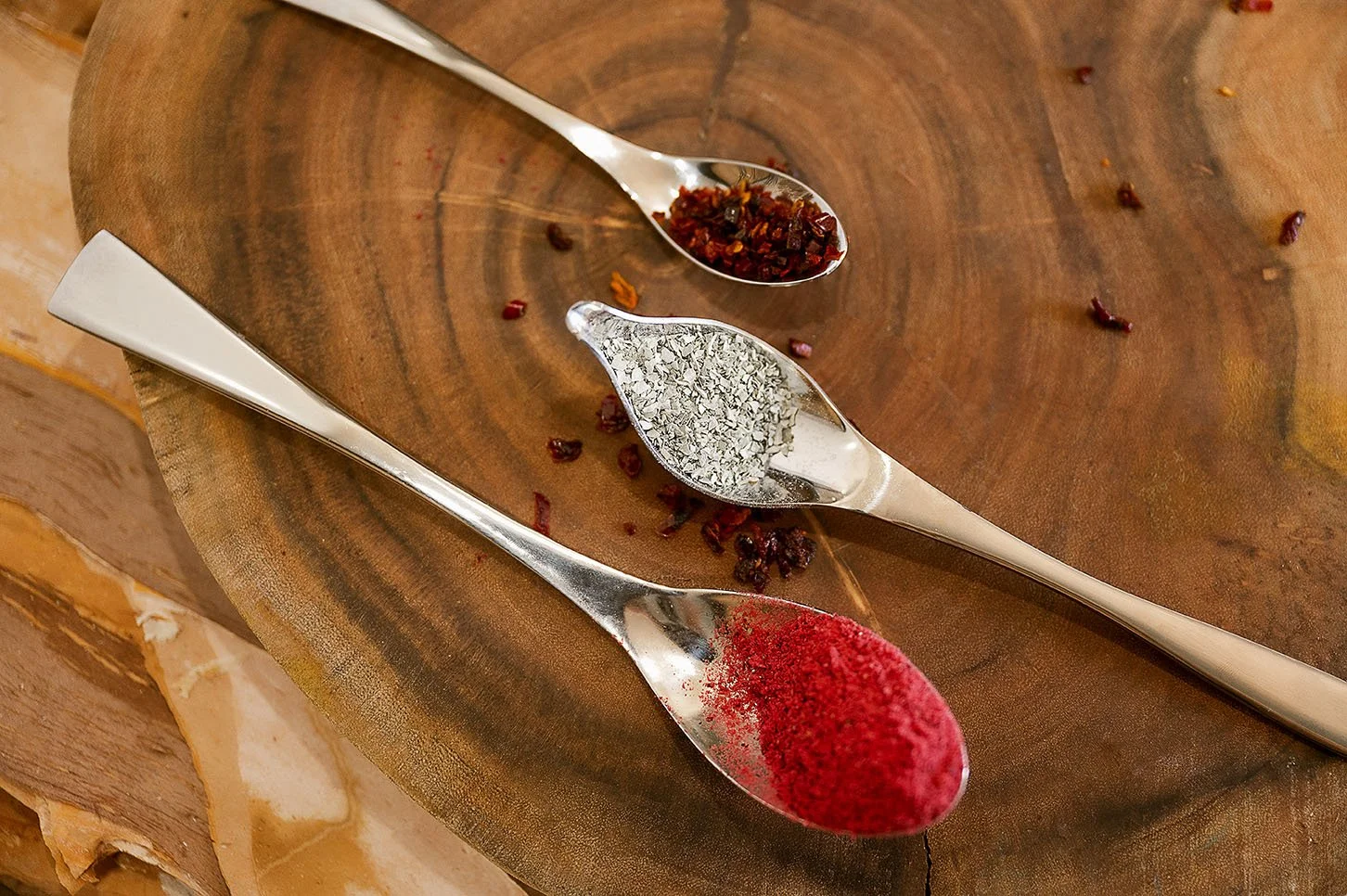 Three metal spoons containing crushed red pepper, dried herbs, and red spice, resting on a round wooden surface.