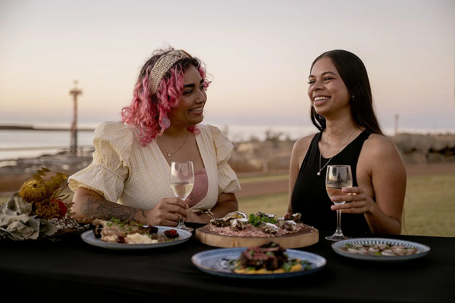 Two women enjoying a meal outdoors at sunset, holding glasses of wine, with plates of food on the table.