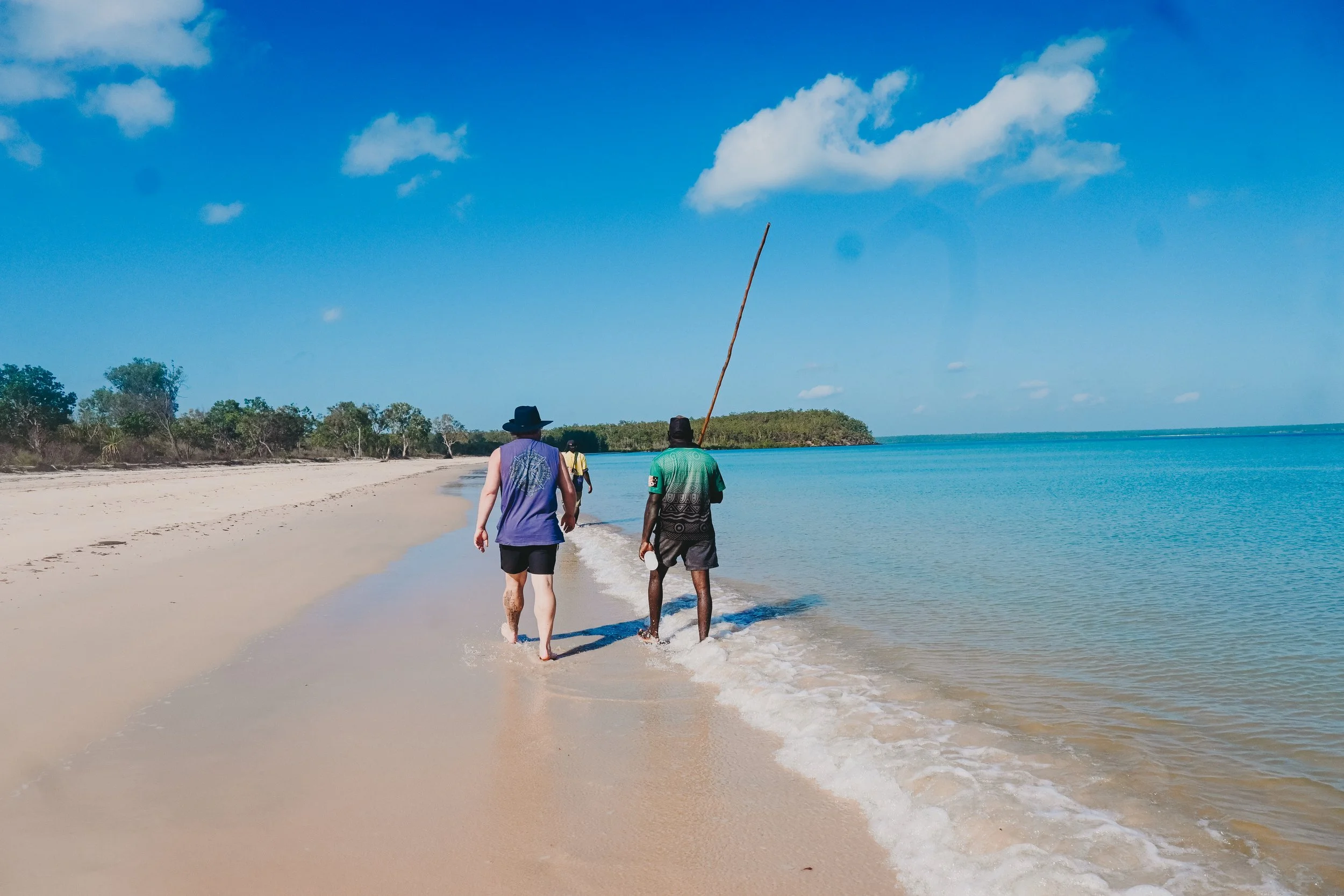 Three people walking along a sandy beach with clear blue water and sky, one carrying a long stick.