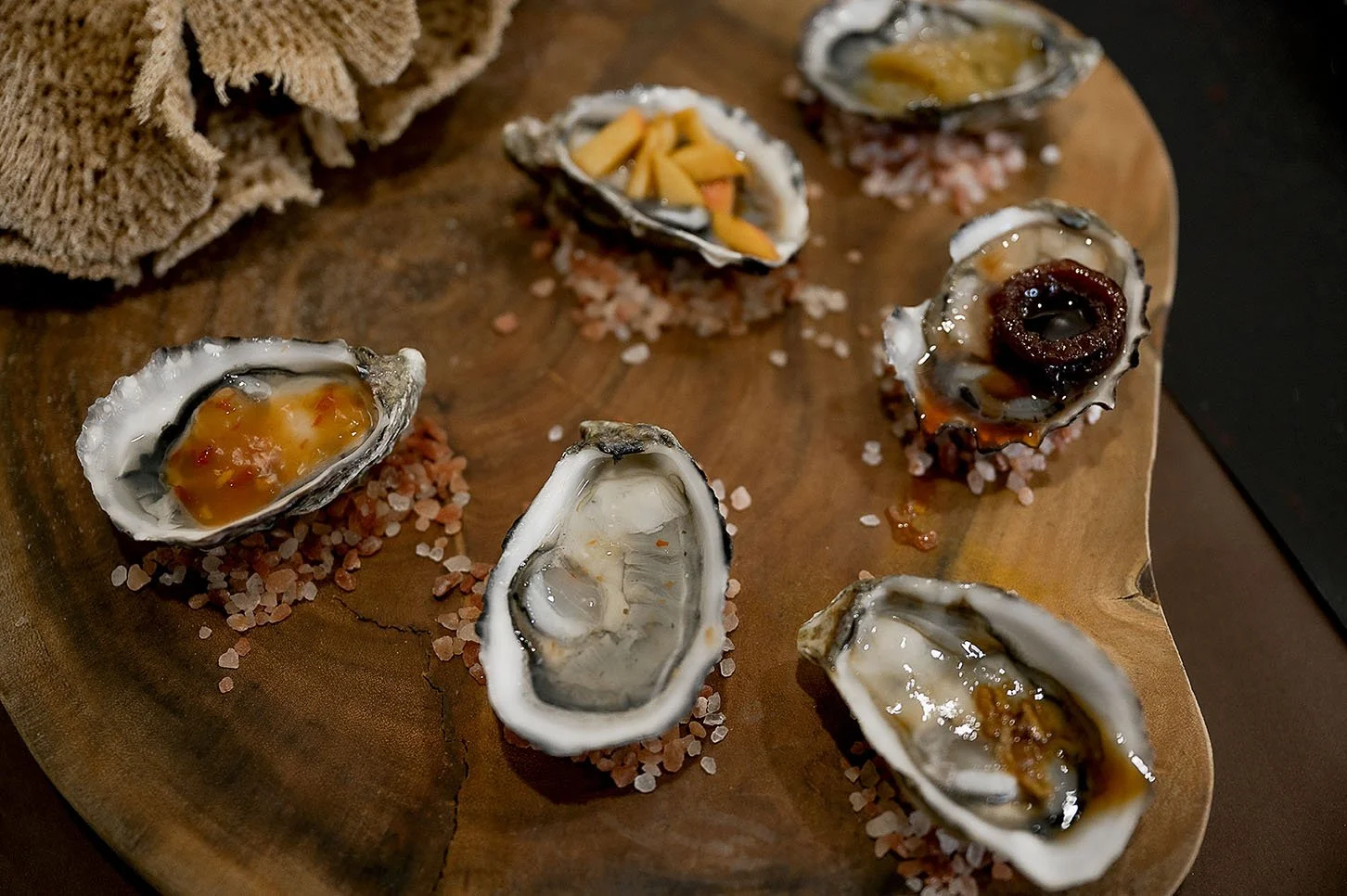 Six raw oysters on the half shell topped with different sauces, served on a wooden platter with Himalayan salt and a cloth napkin.