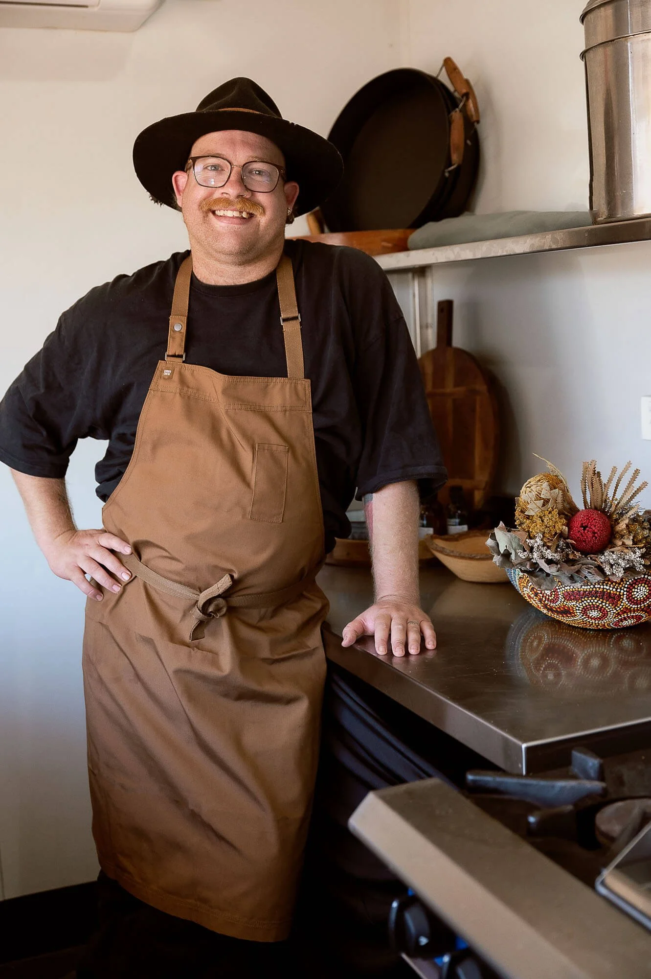 A man wearing glasses, a wide-brimmed hat, a black t-shirt, and a tan apron standing in a kitchen. He is smiling and resting his left hand on a stainless steel countertop, next to a decorative bowl with food ingredients in it.
