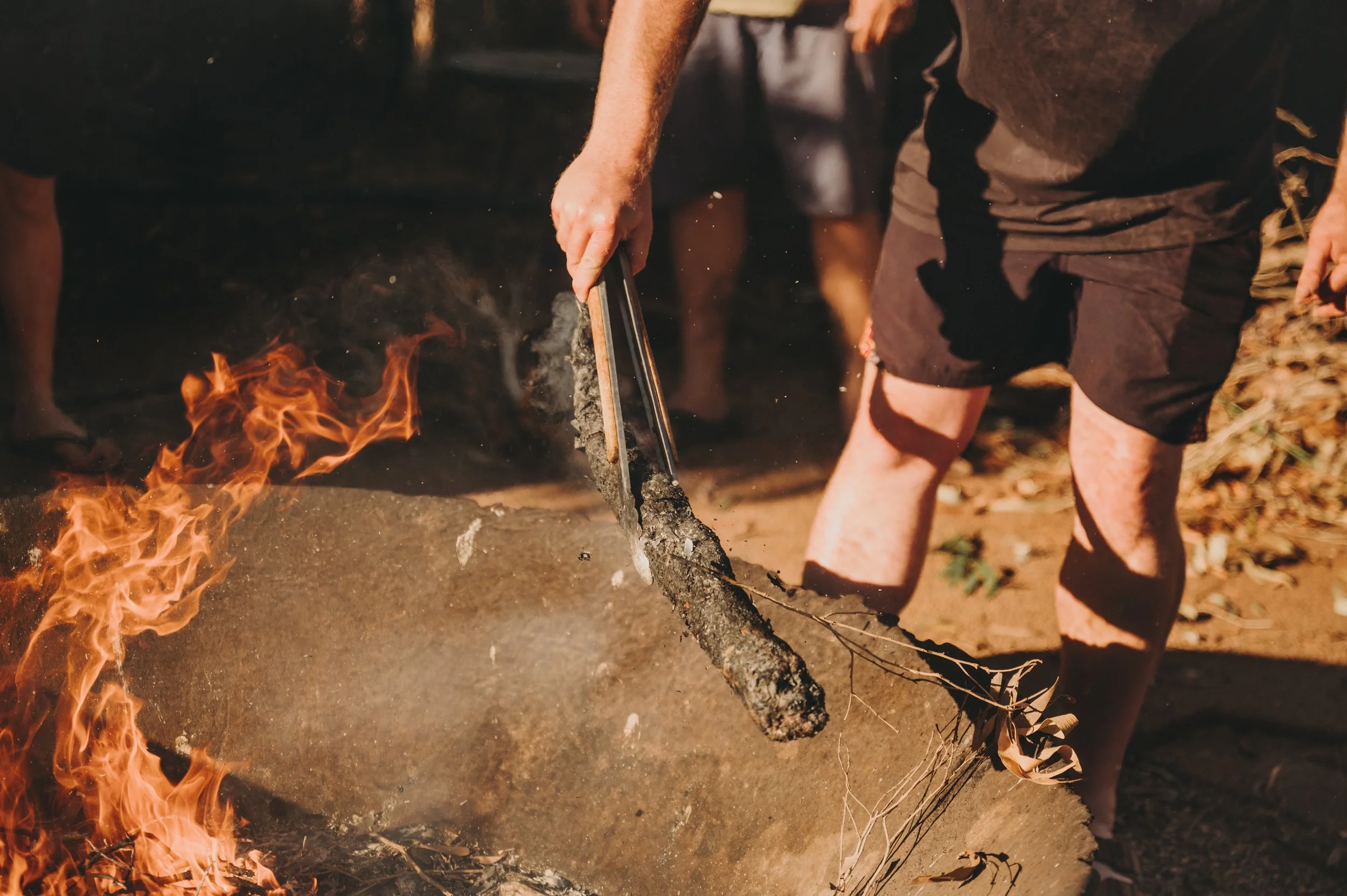 Person in shorts using tongs to burn woody debris in a fire pit surrounded by others.
