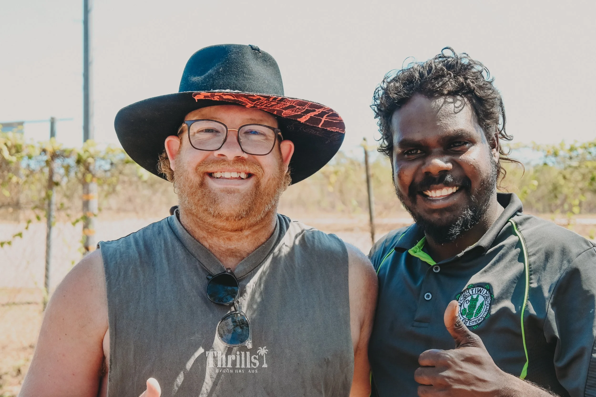 Two smiling men standing outdoors, one wearing a large black hat with red pattern trim, glasses, and a sleeveless gray shirt, and the other with curly hair, a beard, and wearing a black athletic shirt with logo, giving a thumbs up.