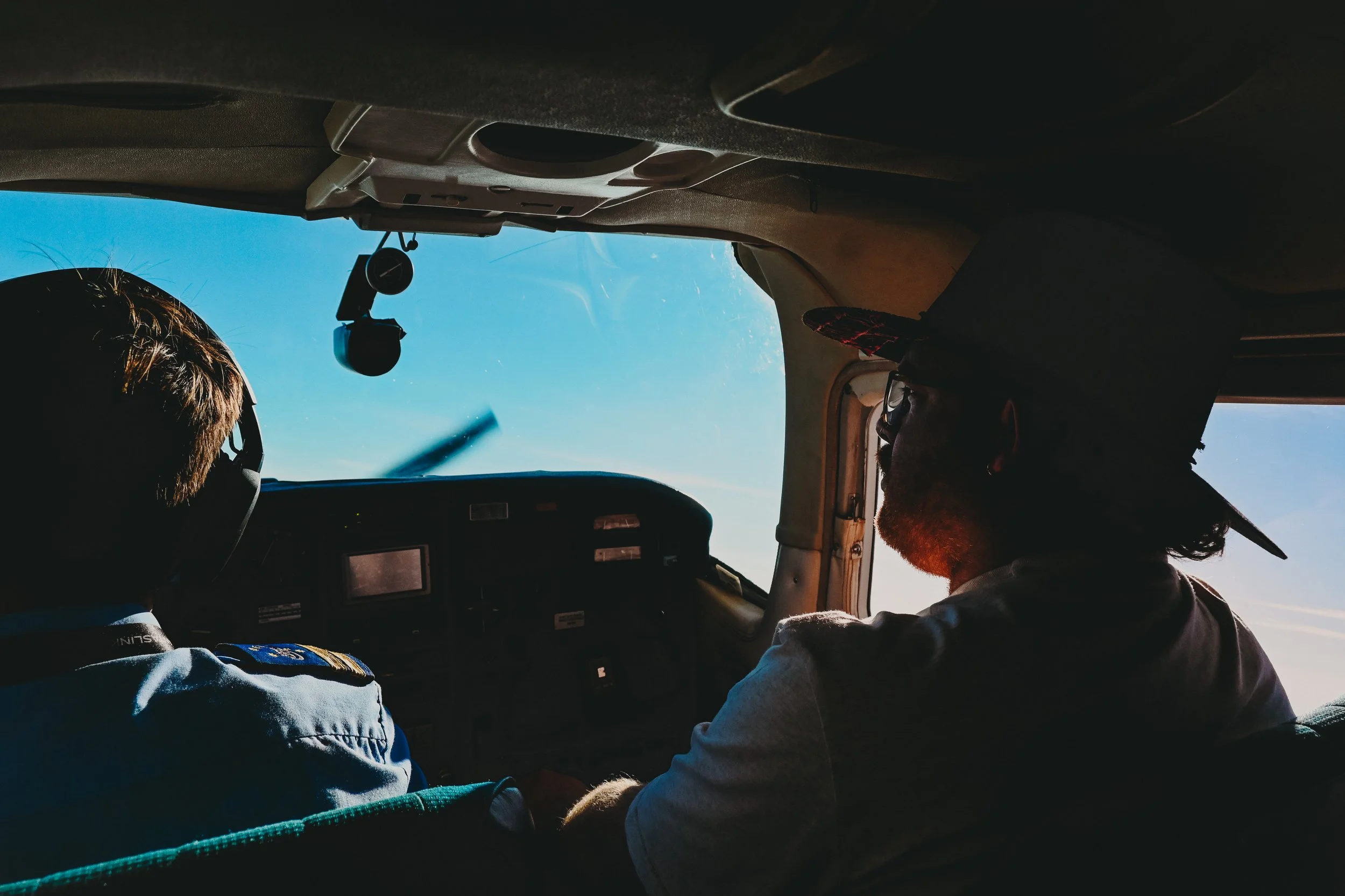 The image shows the interior of an airplane cockpit with two pilots, one wearing a cap and glasses looking towards the sky, and the other in a pilot uniform with a shoulder patch, both seated and facing forward under a clear blue sky.