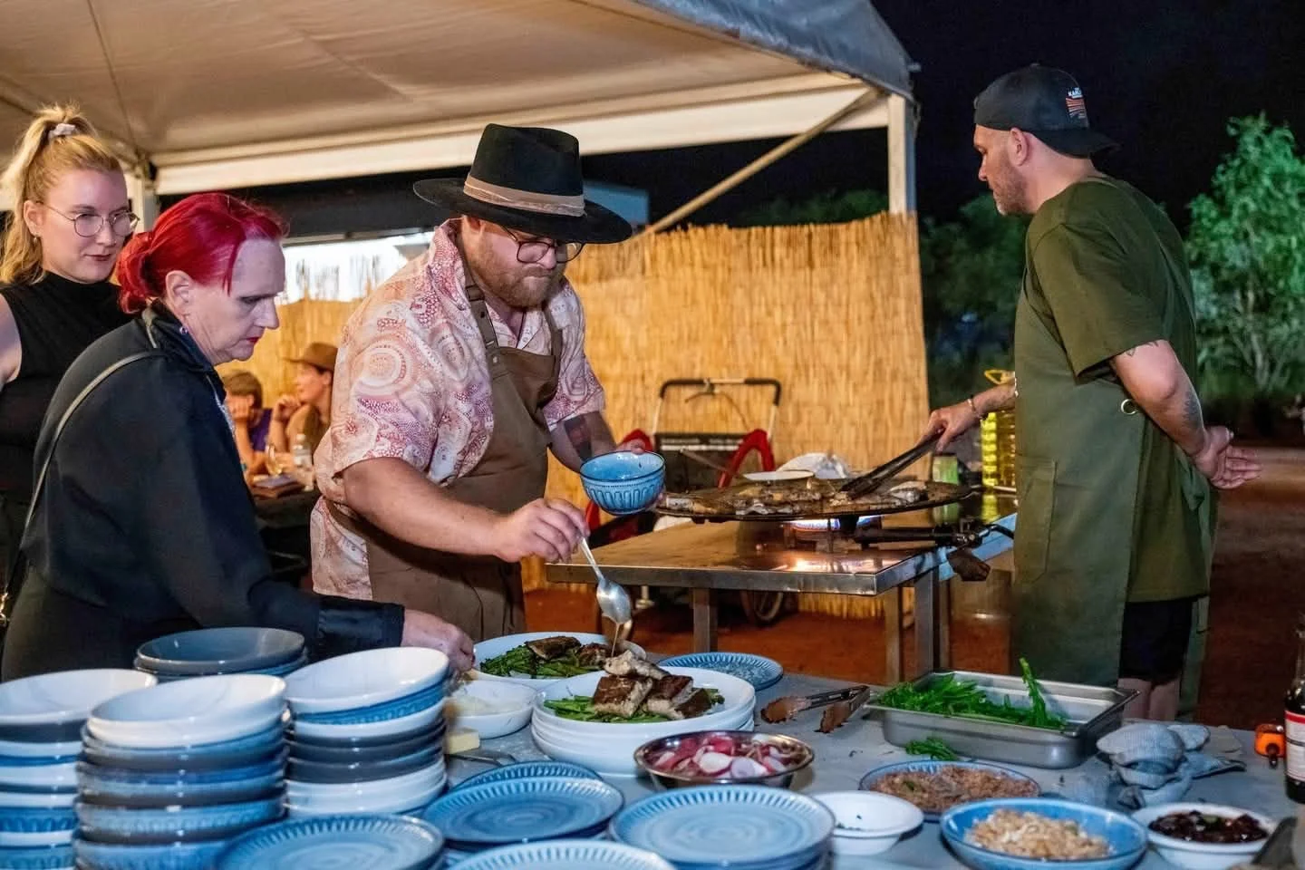 People preparing and serving food at a nighttime outdoor gathering, with a woman and a man cooking on a grill surrounded by dishes and plates.