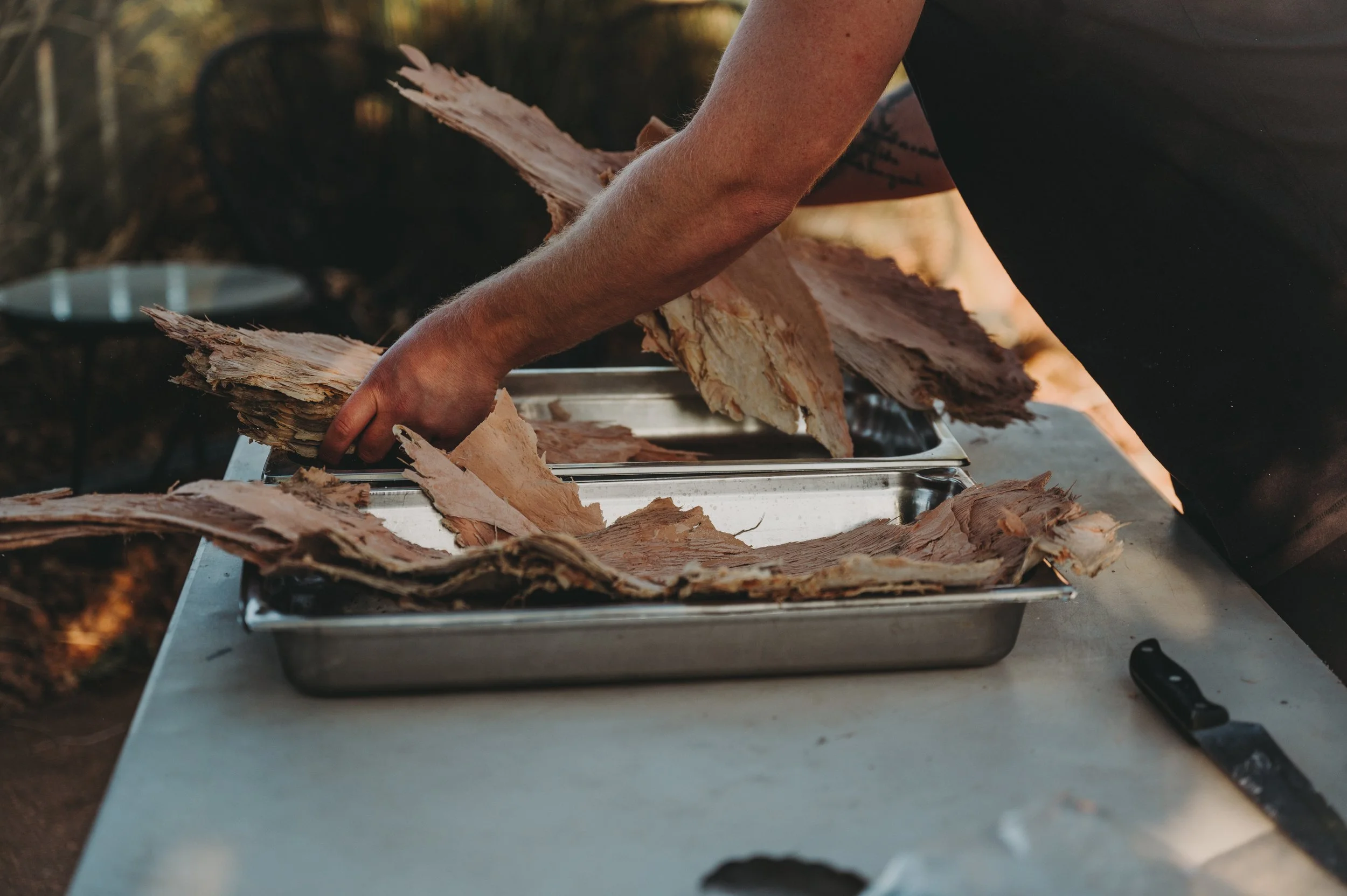 Person handling shredded wood or bark on a metal tray outside.